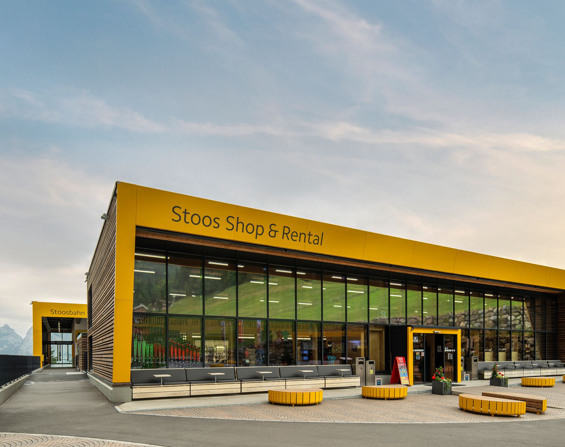Modernes Gebäude des Stoos Shop & Rental mit grosser Glasfassade und gelber Architektur, aufgenommen bei Abendlicht.