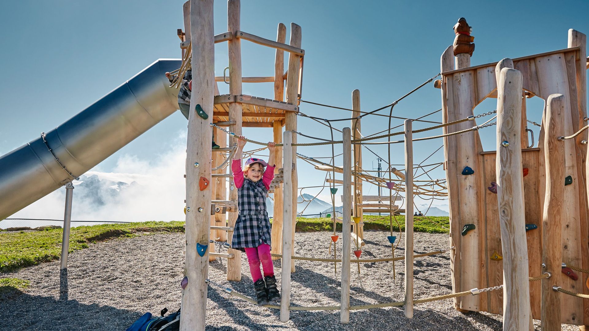 Child climbing on a wooden playground in an alpine setting.
