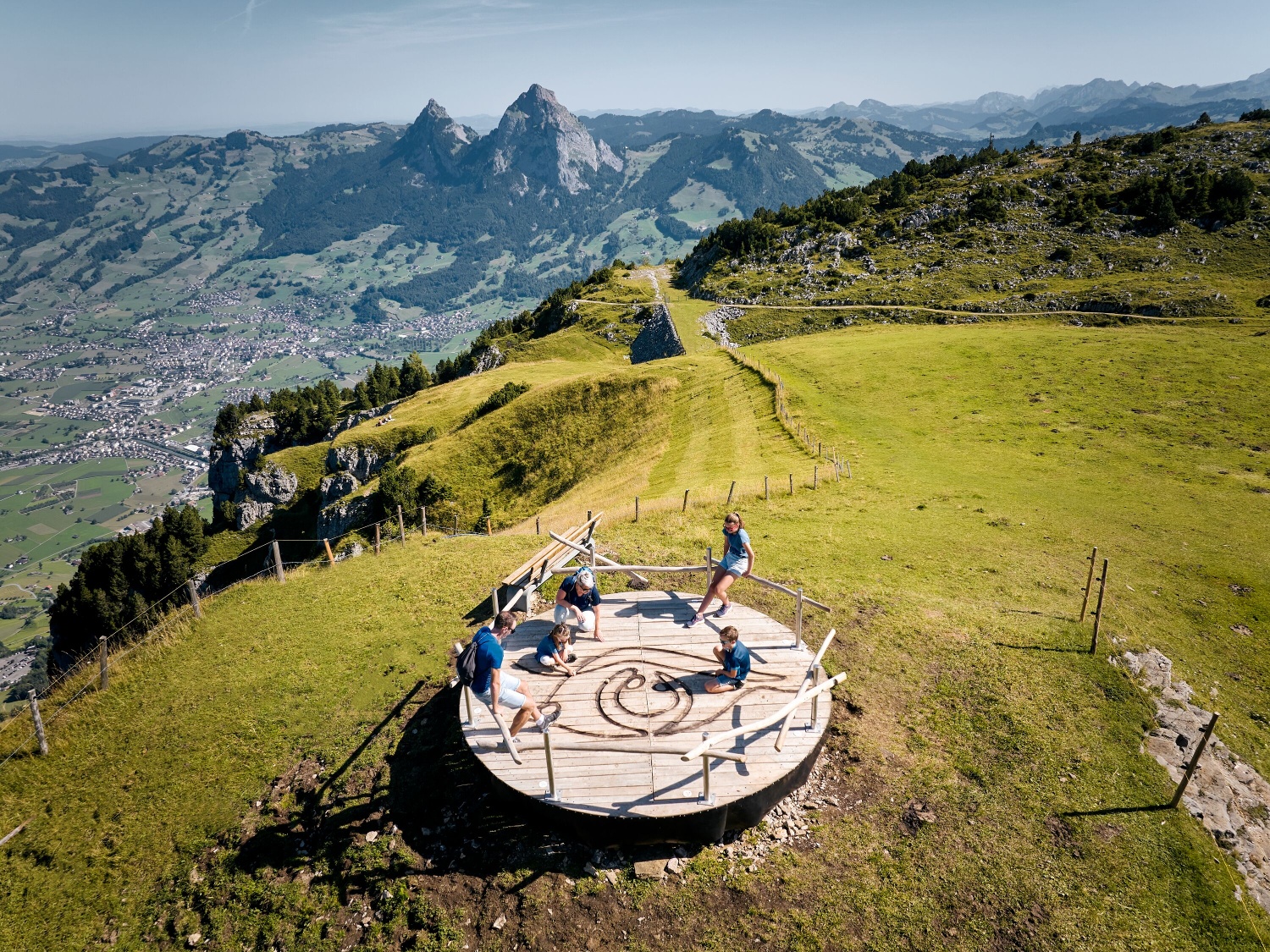 Children play on a round wooden platform on a mountain meadow with views of the Alps.