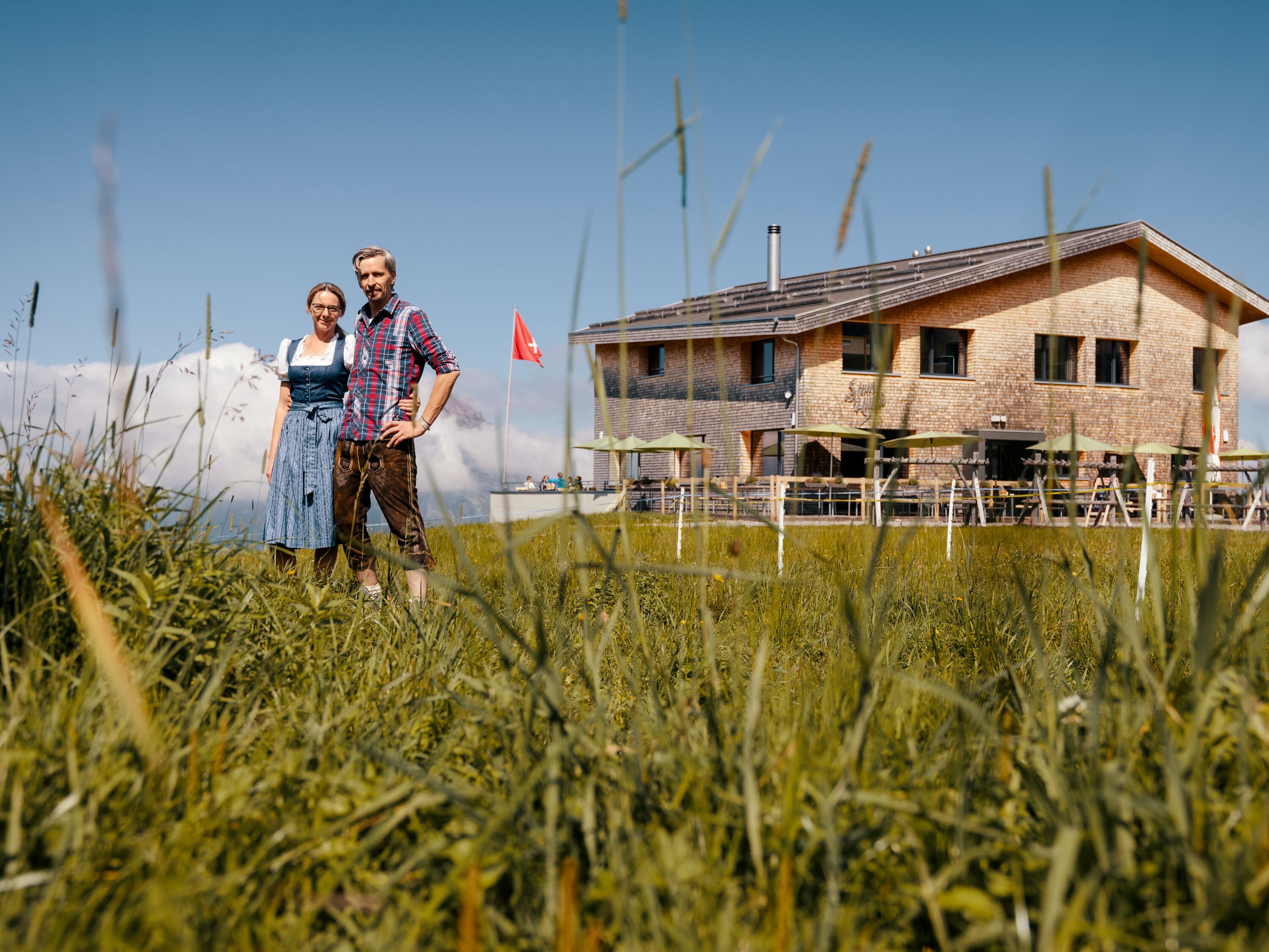 Host couple standing in front of a mountain restaurant on a meadow with views of the alpine landscape.