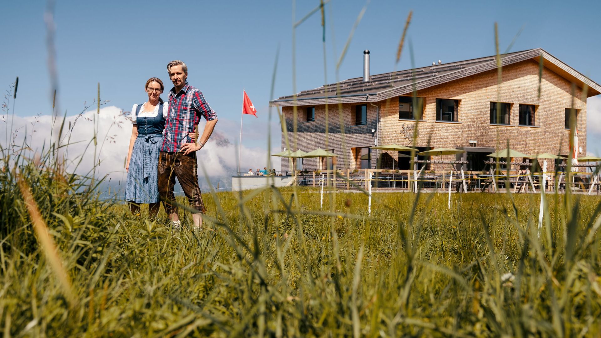Host couple standing in front of a mountain restaurant on a meadow with views of the alpine landscape.