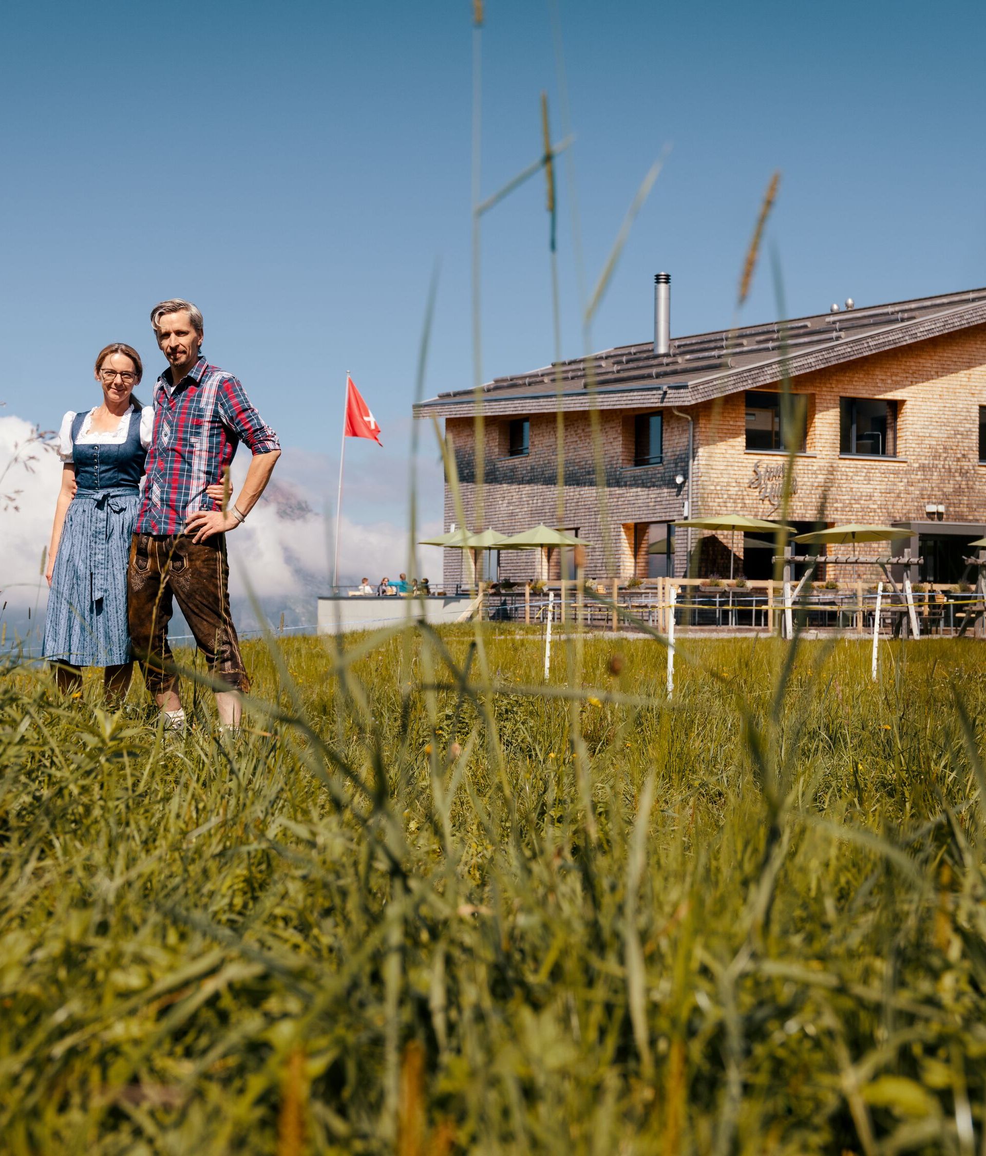 Wirtepaar steht vor einem Bergrestaurant auf einer Wiese mit Blick auf die alpine Landschaft.