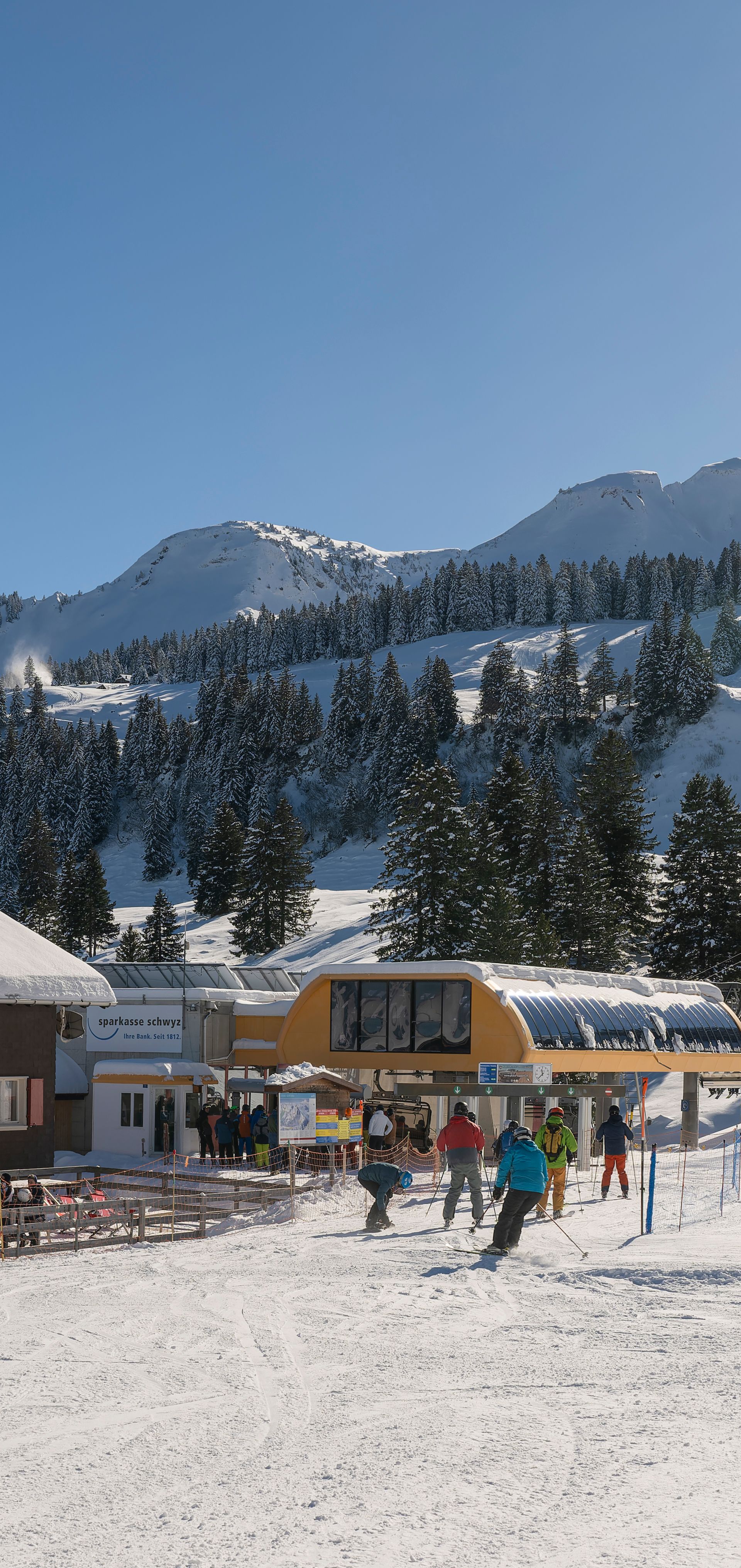 Bergrestaurant an der Skipiste mit Skifahrern und Sesselbahn in verschneiter Berglandschaft.
