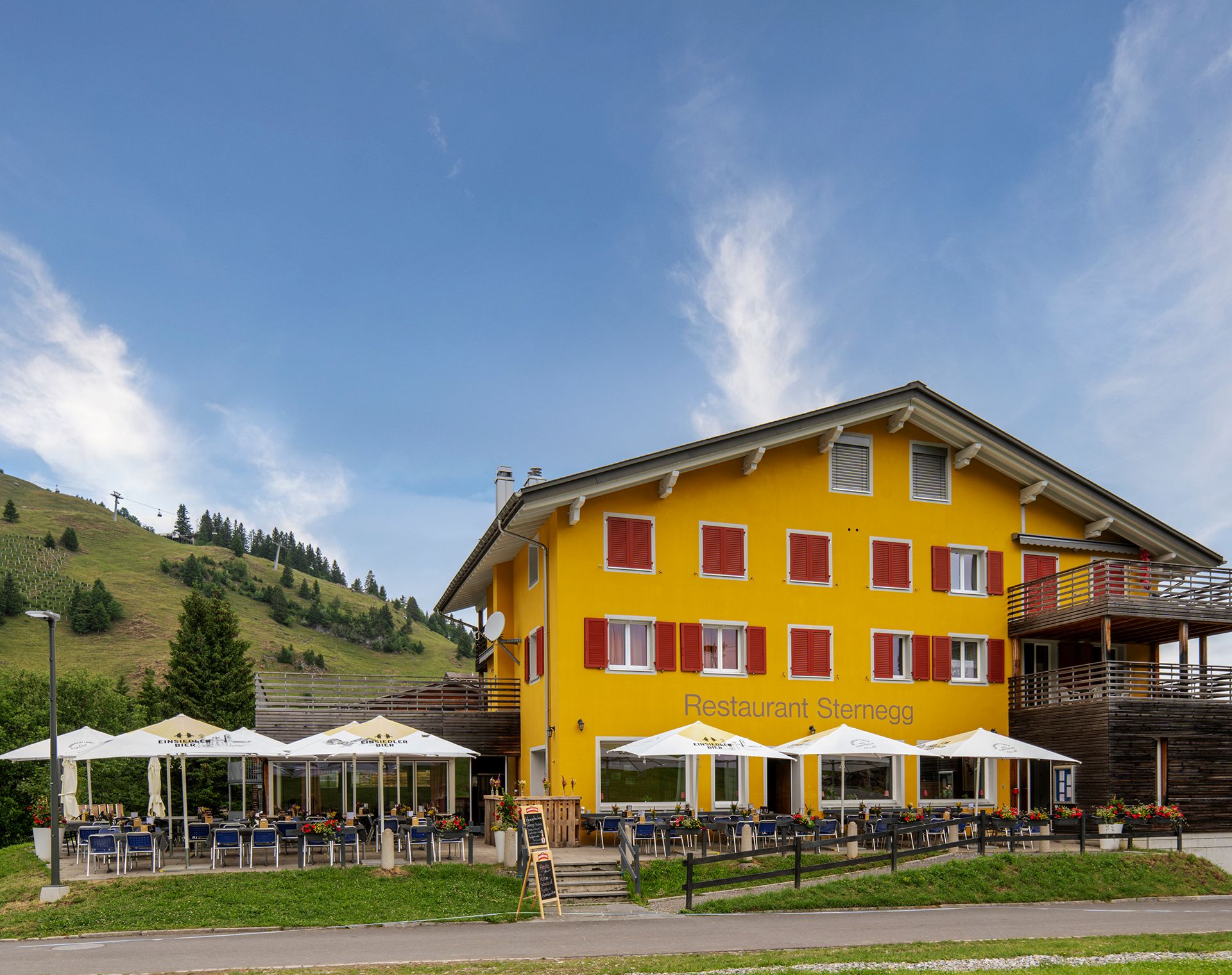 Gelbes Bergrestaurant mit Terrasse auf einer Wiese in alpiner Landschaft.