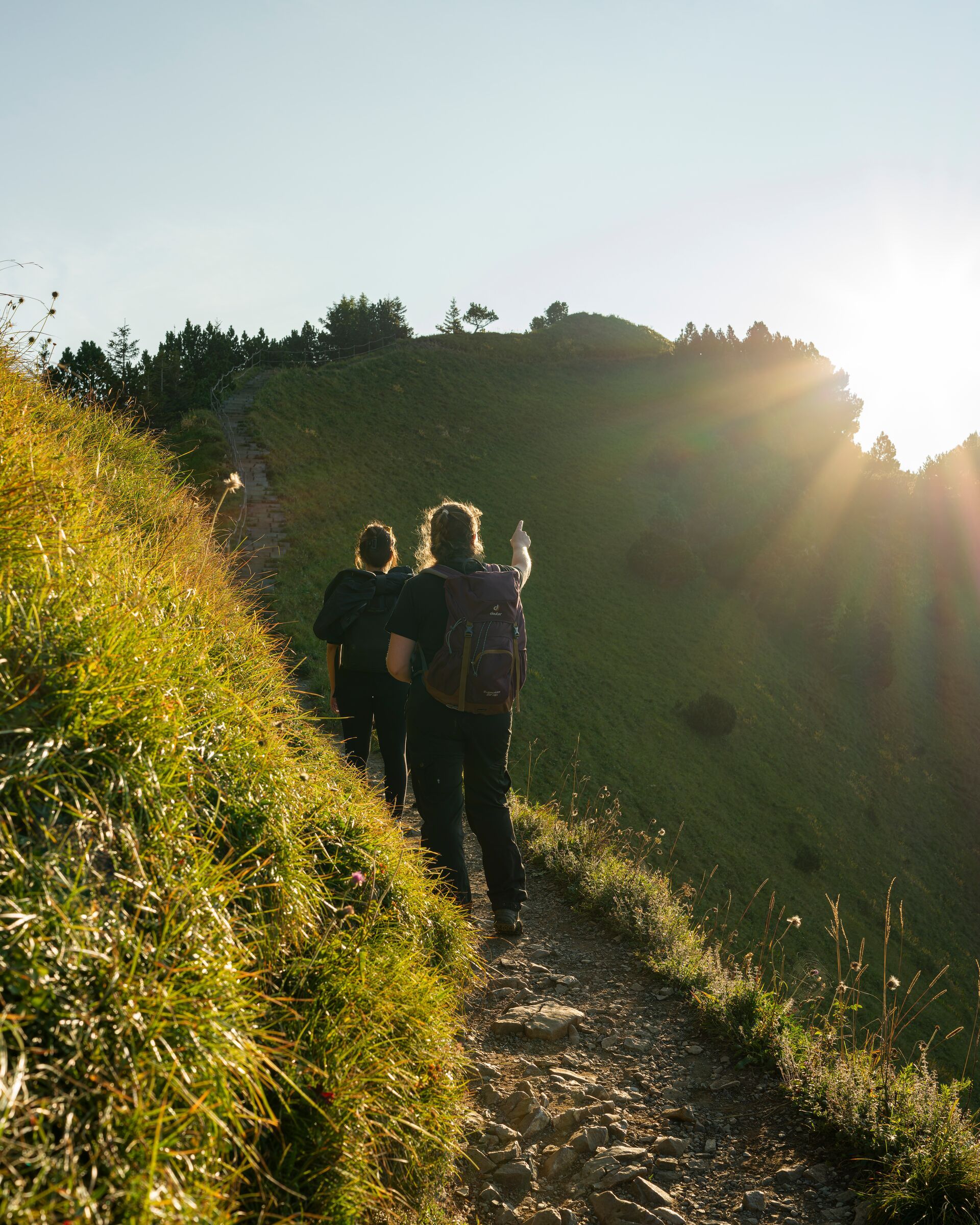Zwei Wanderer auf einem schmalen Bergweg im Gegenlicht der Sonne, umgeben von grünen Hängen.