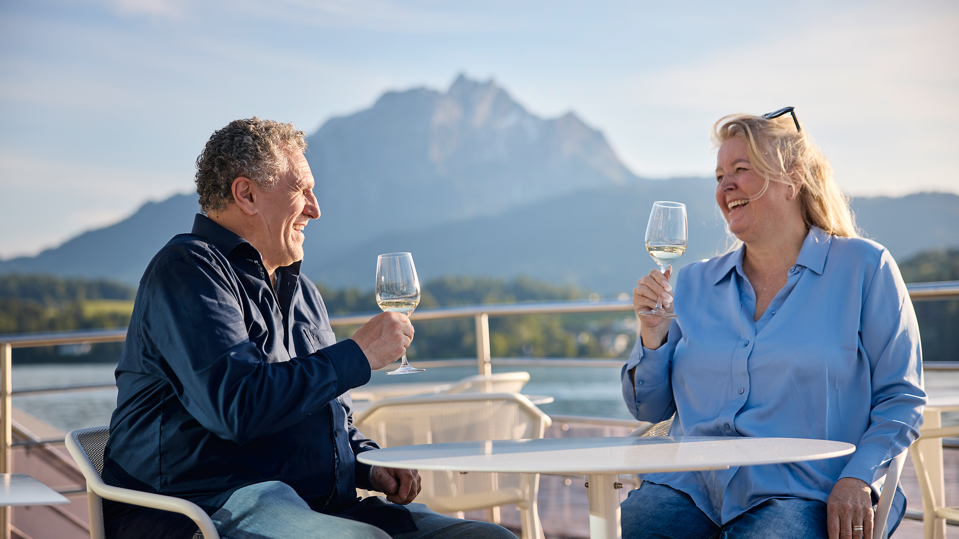 Paar mit Weingläsern an Deck eines Schiffes mit Blick auf den Vierwaldstättersee bei Abendlicht.