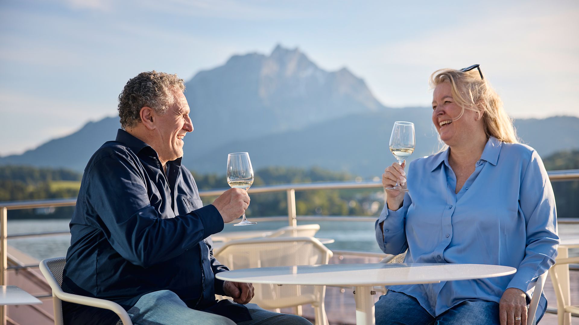 Couple holding wine glasses on the deck of a ship overlooking Lake Lucerne at sunset.