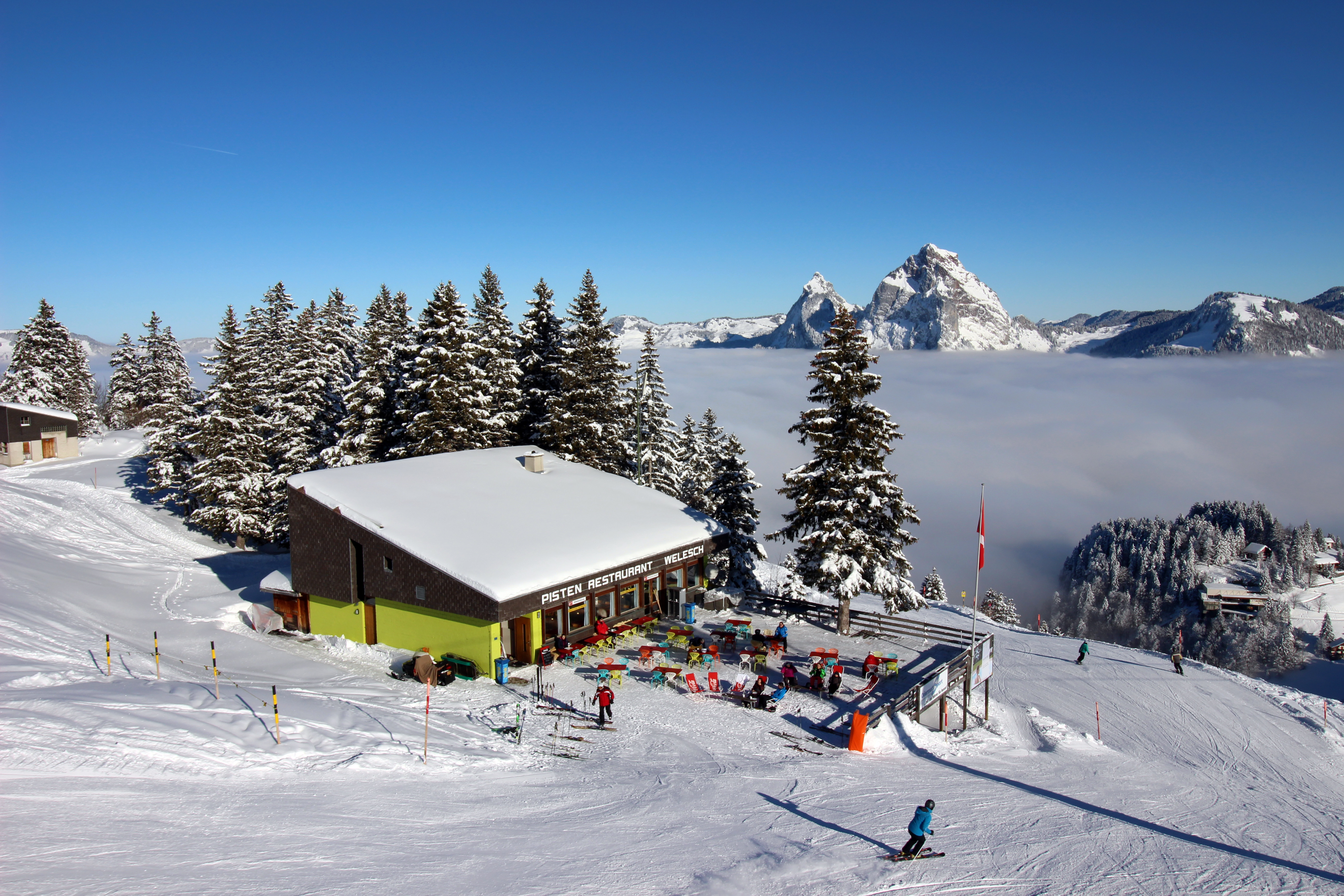 Bergrestaurant an der Skipiste mit Terrasse, Skifahrern und Blick auf verschneite Berge über dem Nebelmeer.