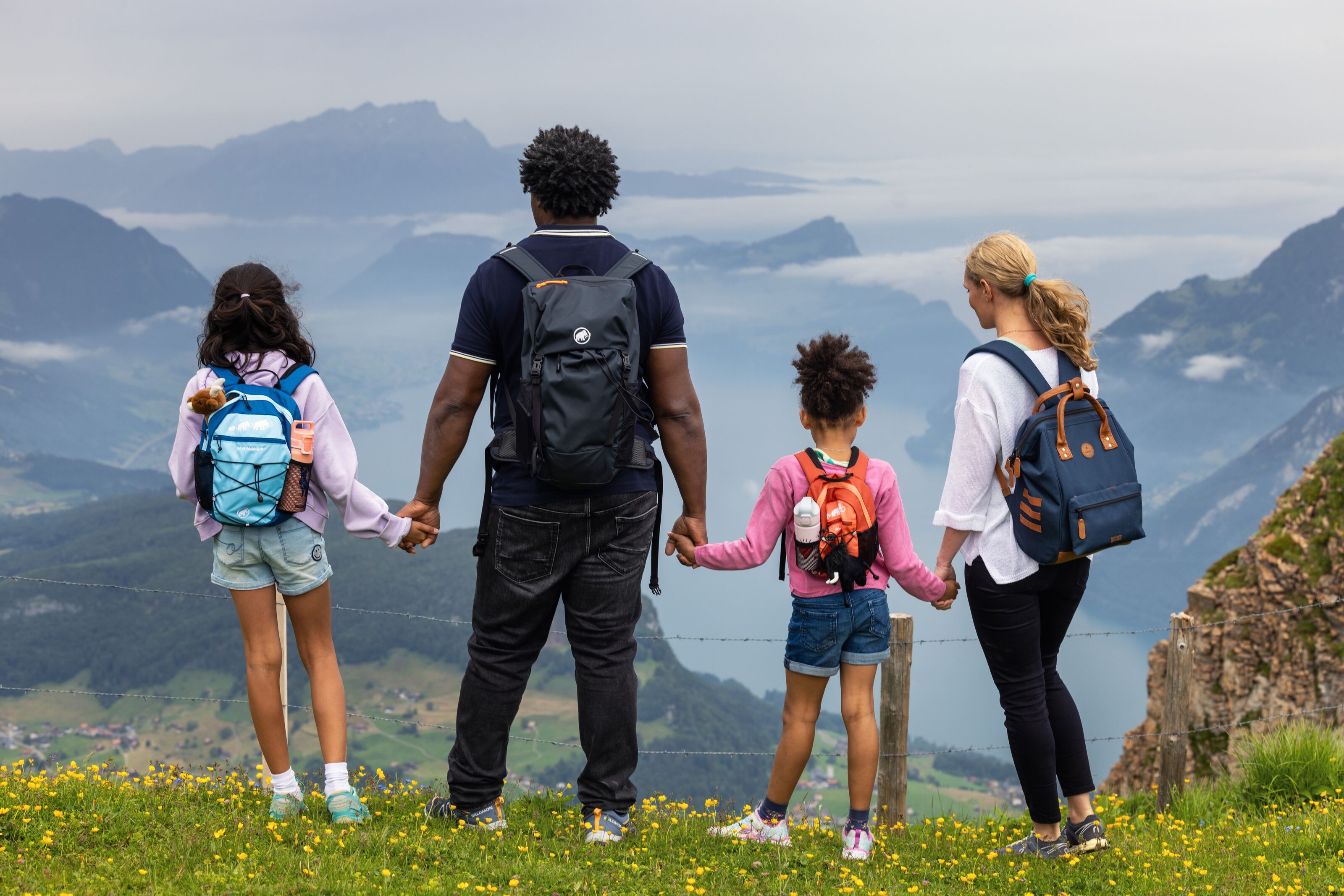 Familie steht auf einer Bergwiese und blickt gemeinsam über eine weite Berg- und Seelandschaft.
