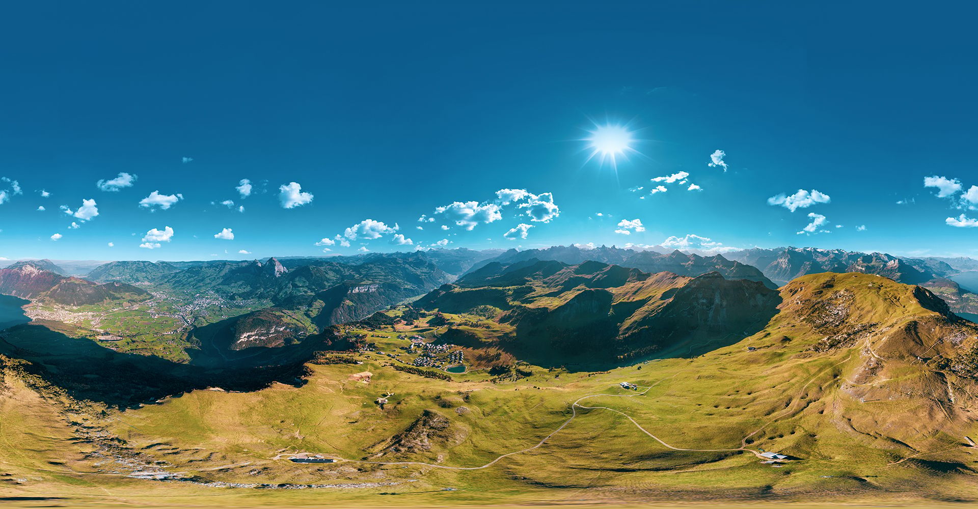 Panoramaaufnahme der Stoos-Region mit weiten alpinen Wiesen, Bergketten und strahlendem Sonnenschein.