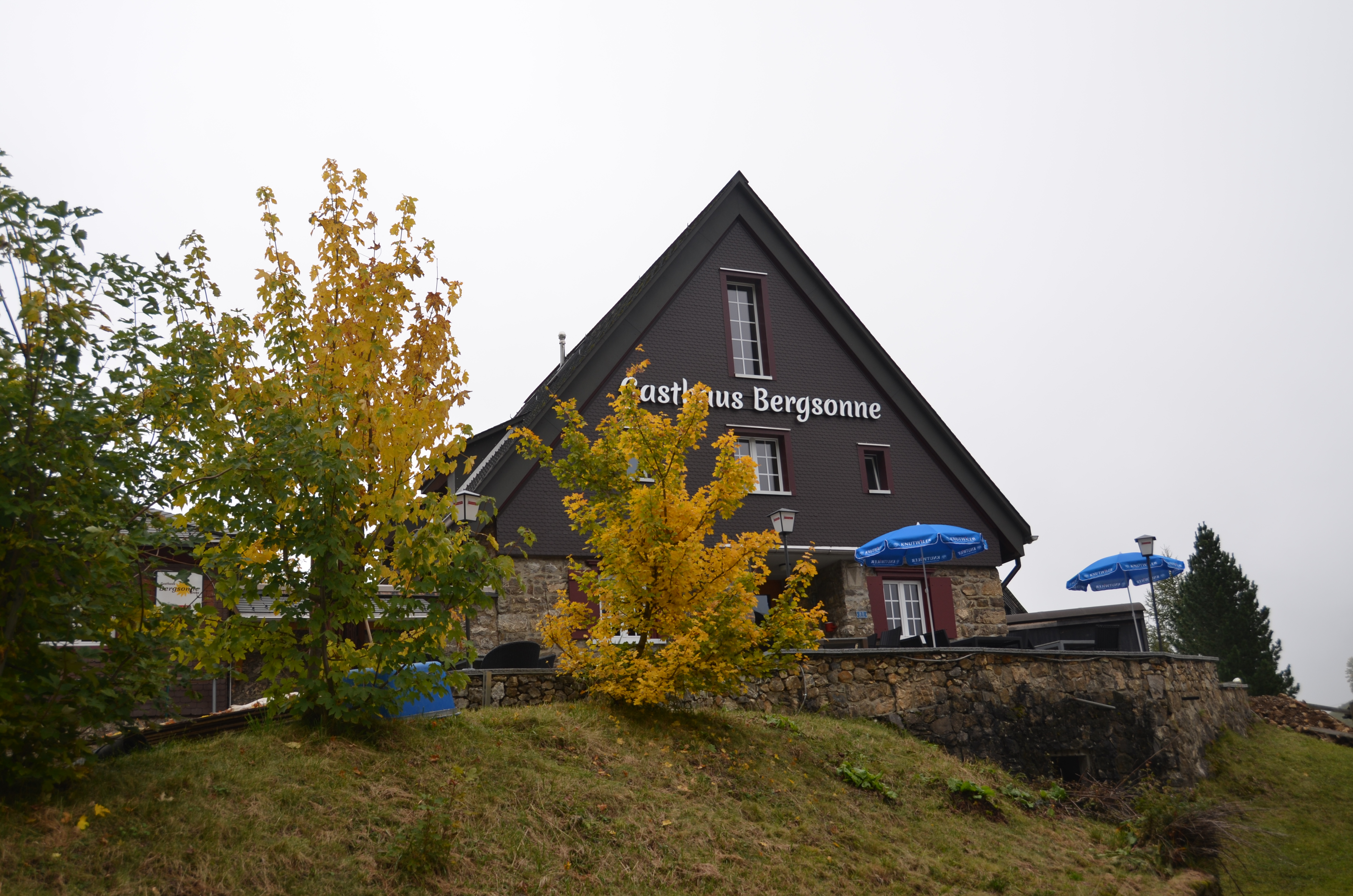 Bergrestaurant mit Terrasse auf einem Hügel in herbstlicher alpiner Landschaft.