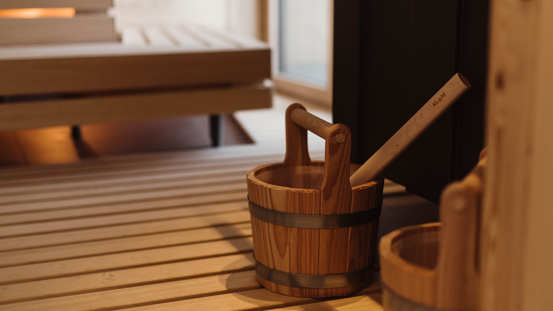 Sauna-Detail mit Holzeimer, Kelle und warm beleuchteten Sitzbänken.
