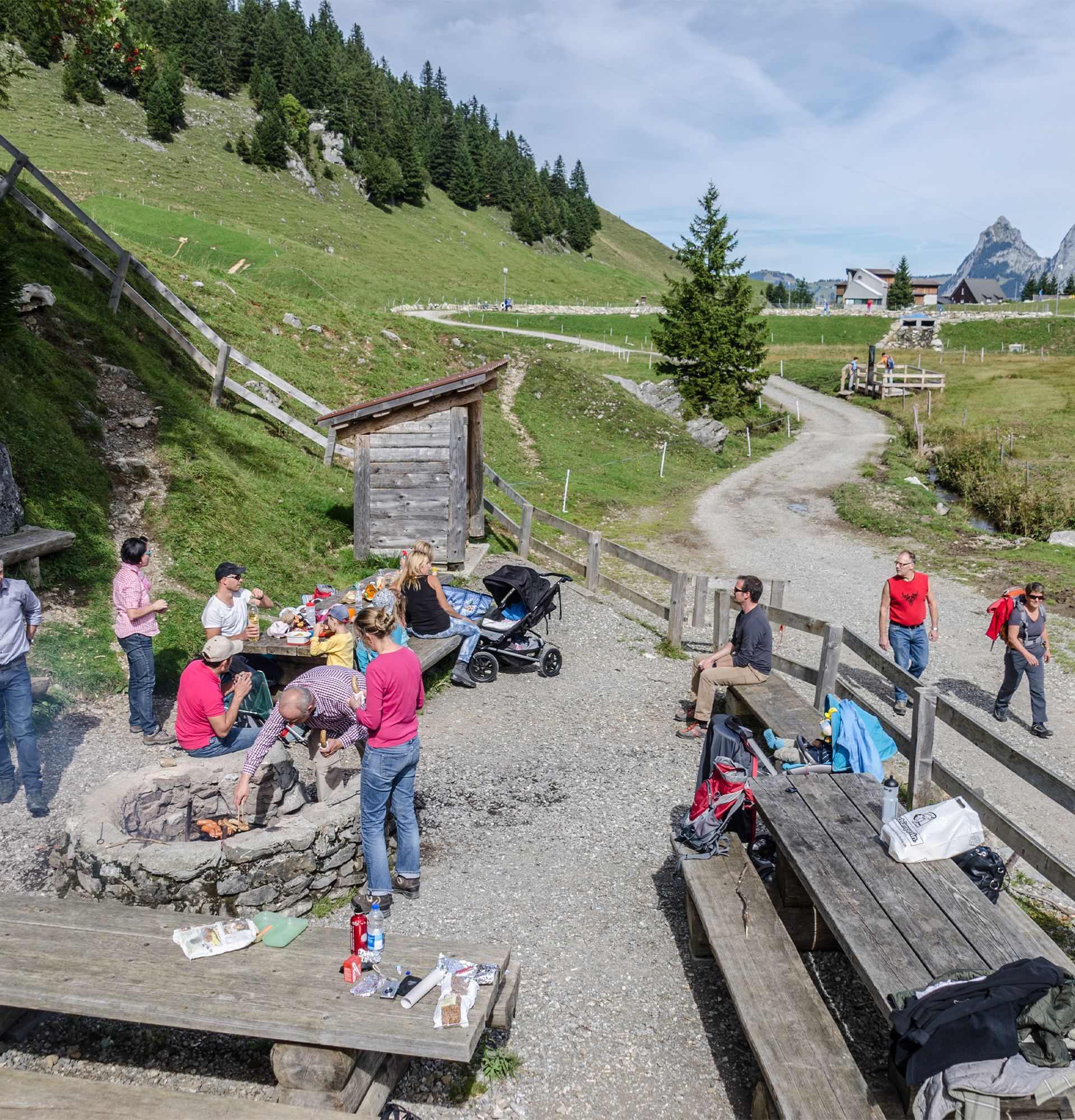 Familien und Kinder auf einem Spiel- und Rastplatz in alpiner Landschaft mit Sitzgelegenheiten und Spielgeräten.
