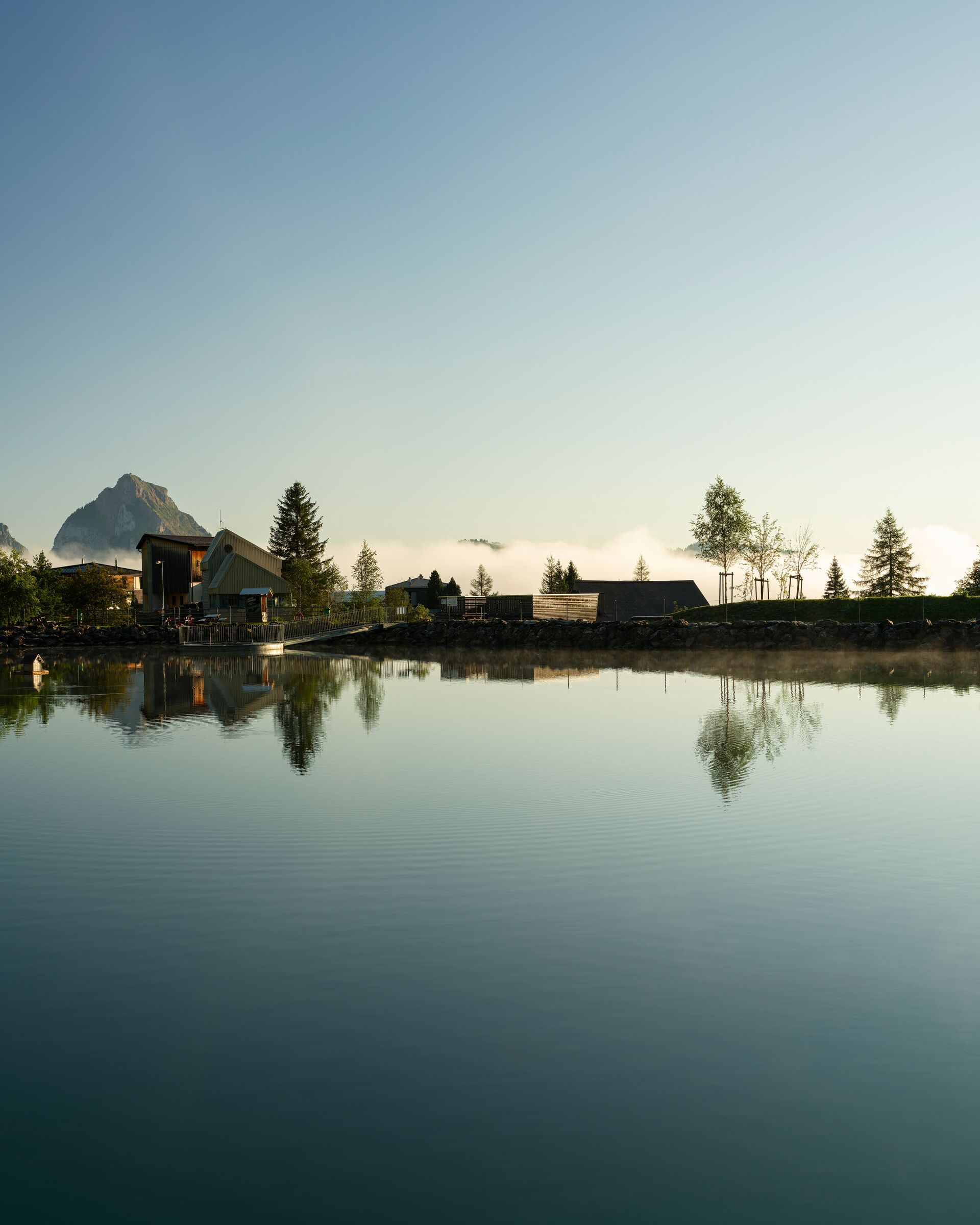 Mountain lake with a calm surface reflecting houses and trees in an alpine landscape.