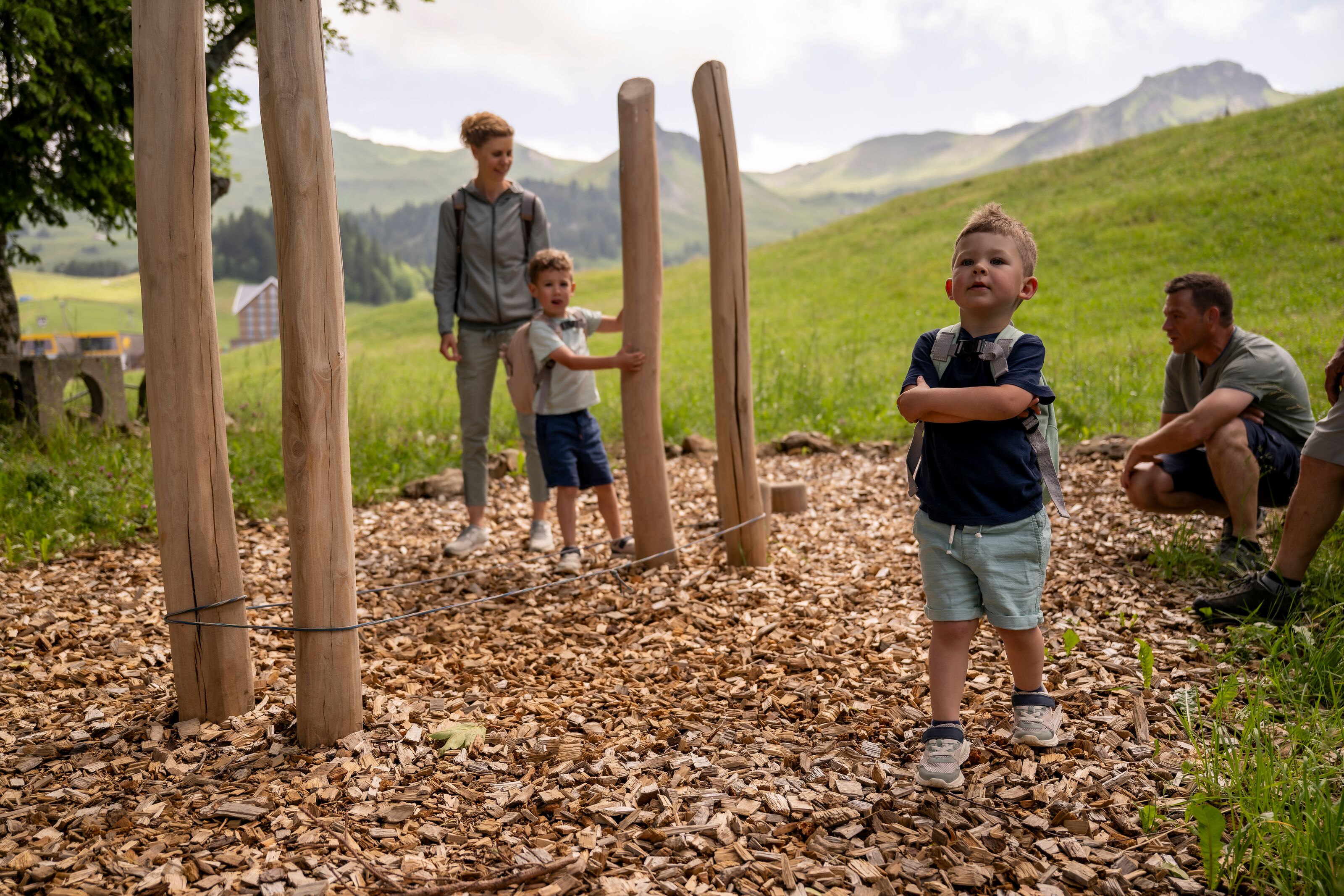 Kinder spielen auf einem Naturspielplatz mit Holzpfosten und Schnitzelboden, während Erwachsene zuschauen.