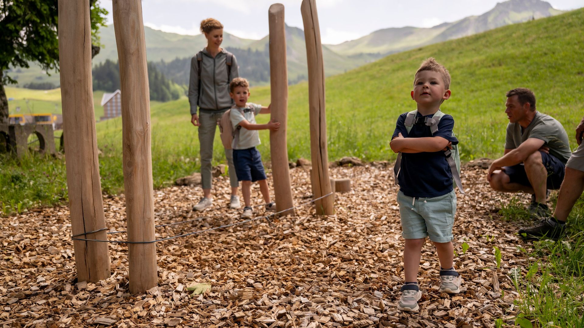 Kinder spielen auf einem Naturspielplatz mit Holzpfosten und Schnitzelboden, während Erwachsene zuschauen.