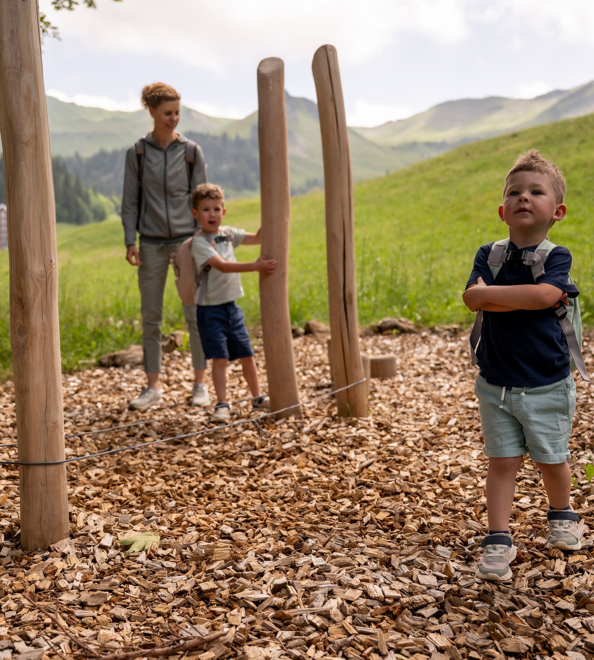 Children playing at a natural playground with wooden posts and woodchip ground while adults watch