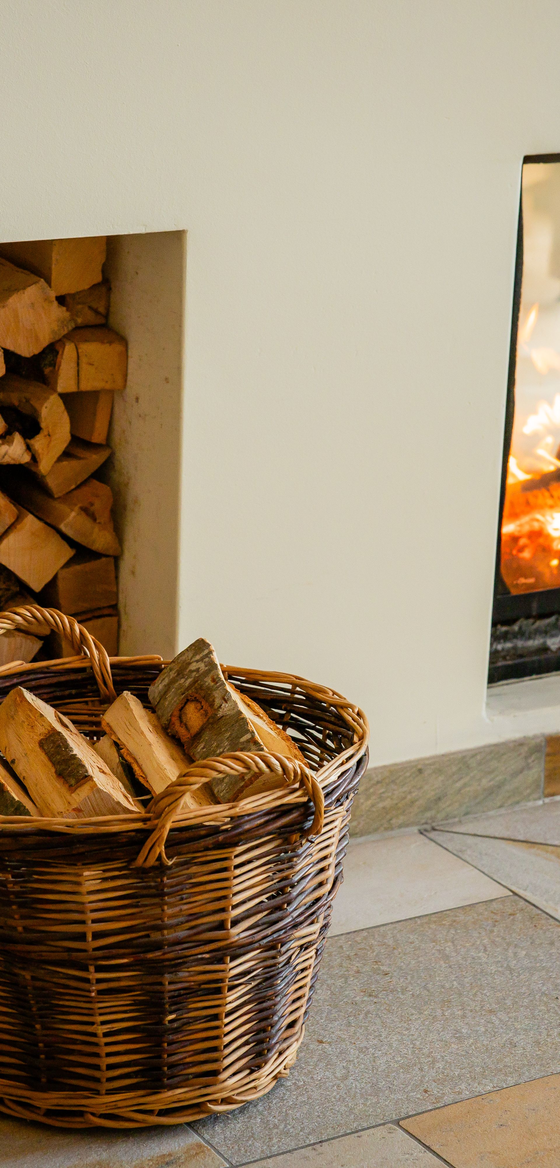 Kamin mit brennendem Feuer neben gestapeltem Holz und Korb in gemütlichem Innenraum.