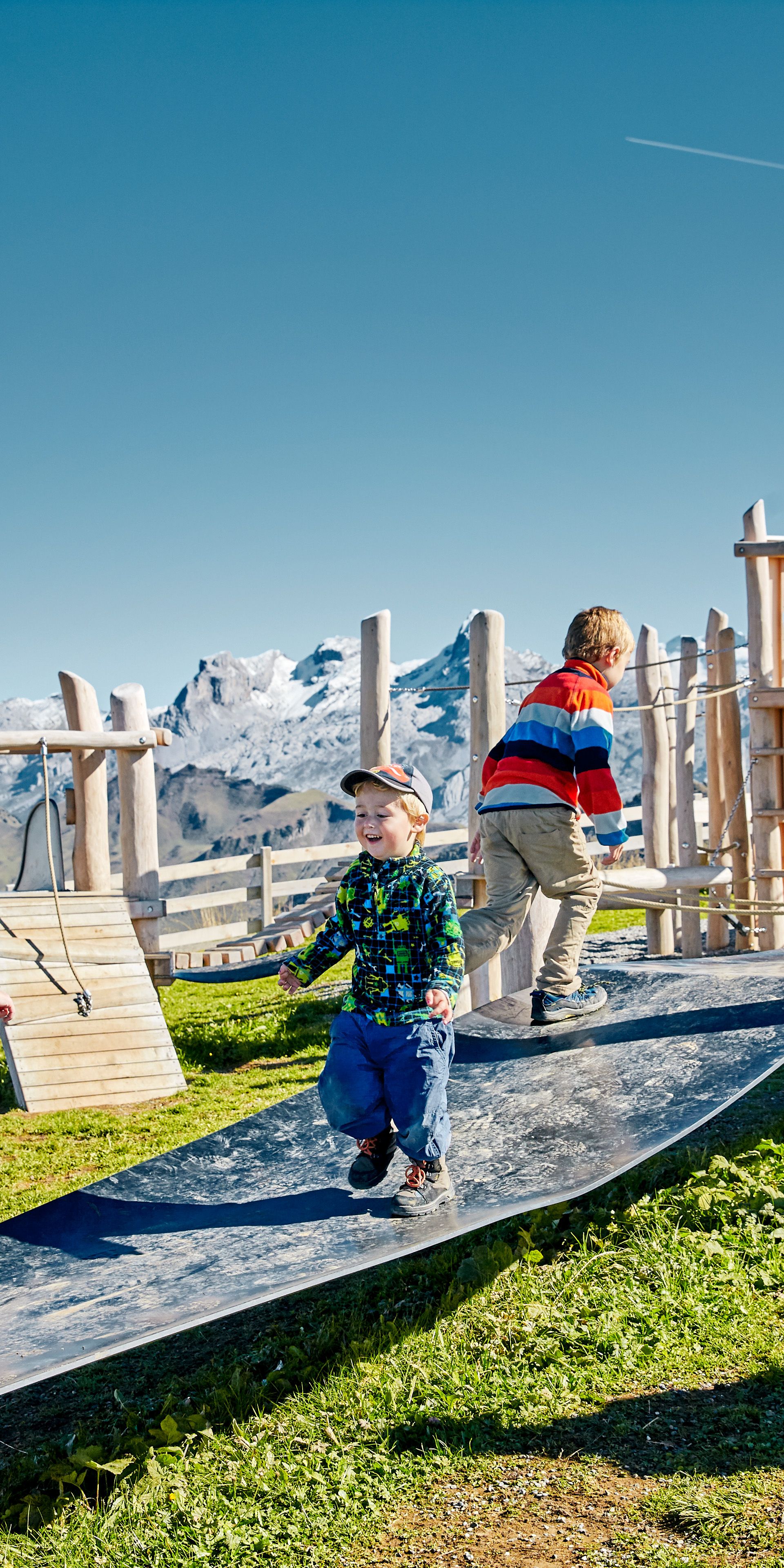 Kinder rennen über einen Spielplatz auf einer Bergwiese mit Blick auf die Alpen.