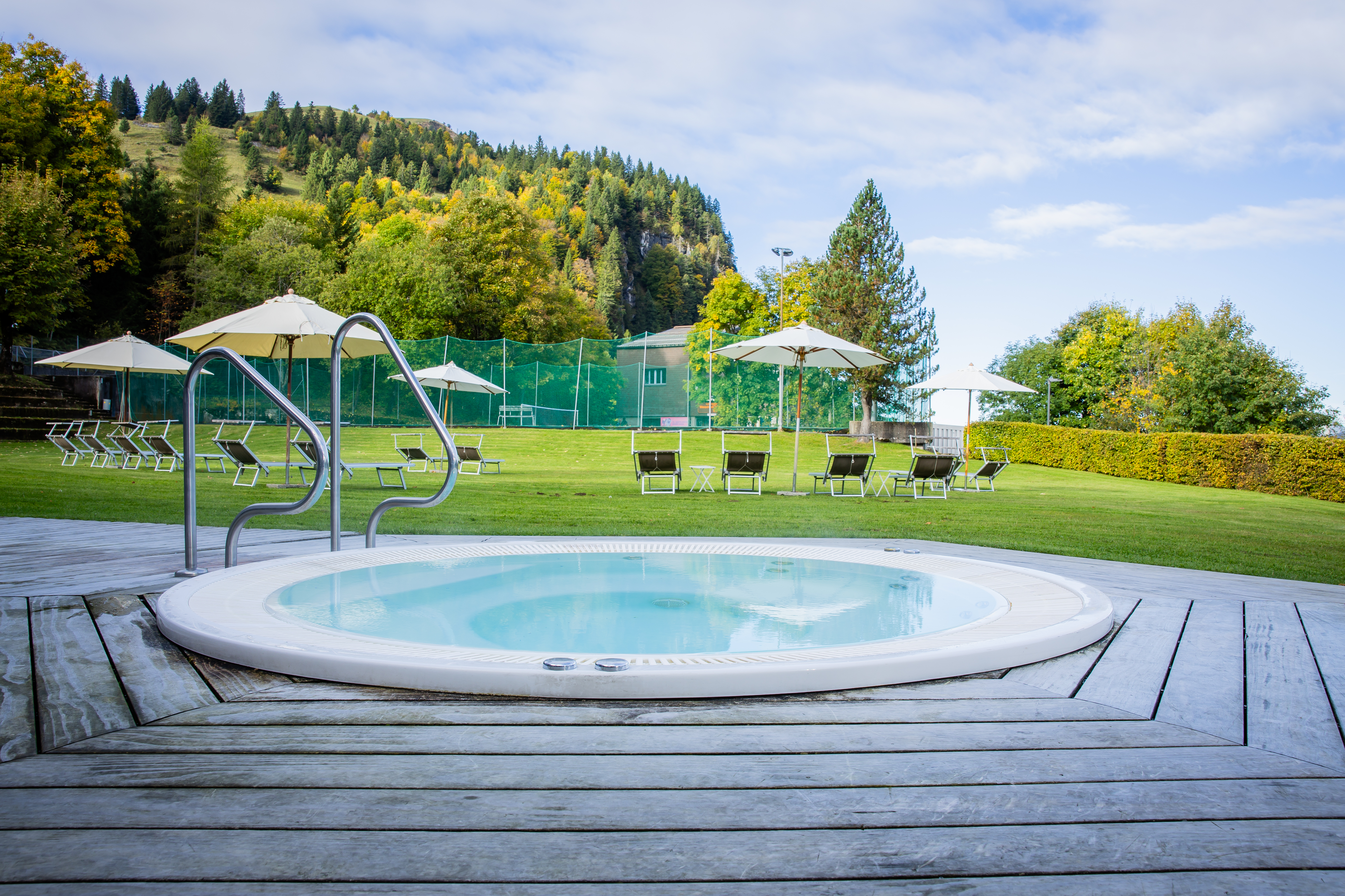 Round outdoor hot tub on a wooden deck with a view of green meadows and trees.