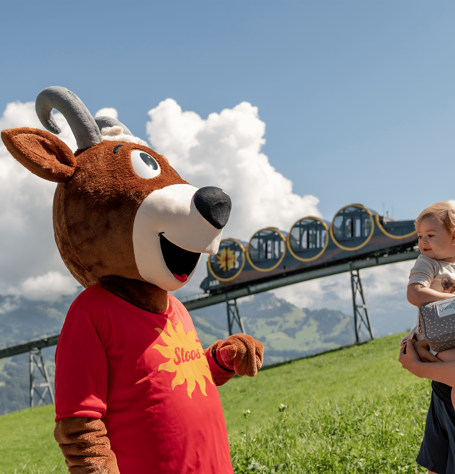 Mascot and child stand on a mountain meadow with views of the Stoos funicular and the alpine landscape.