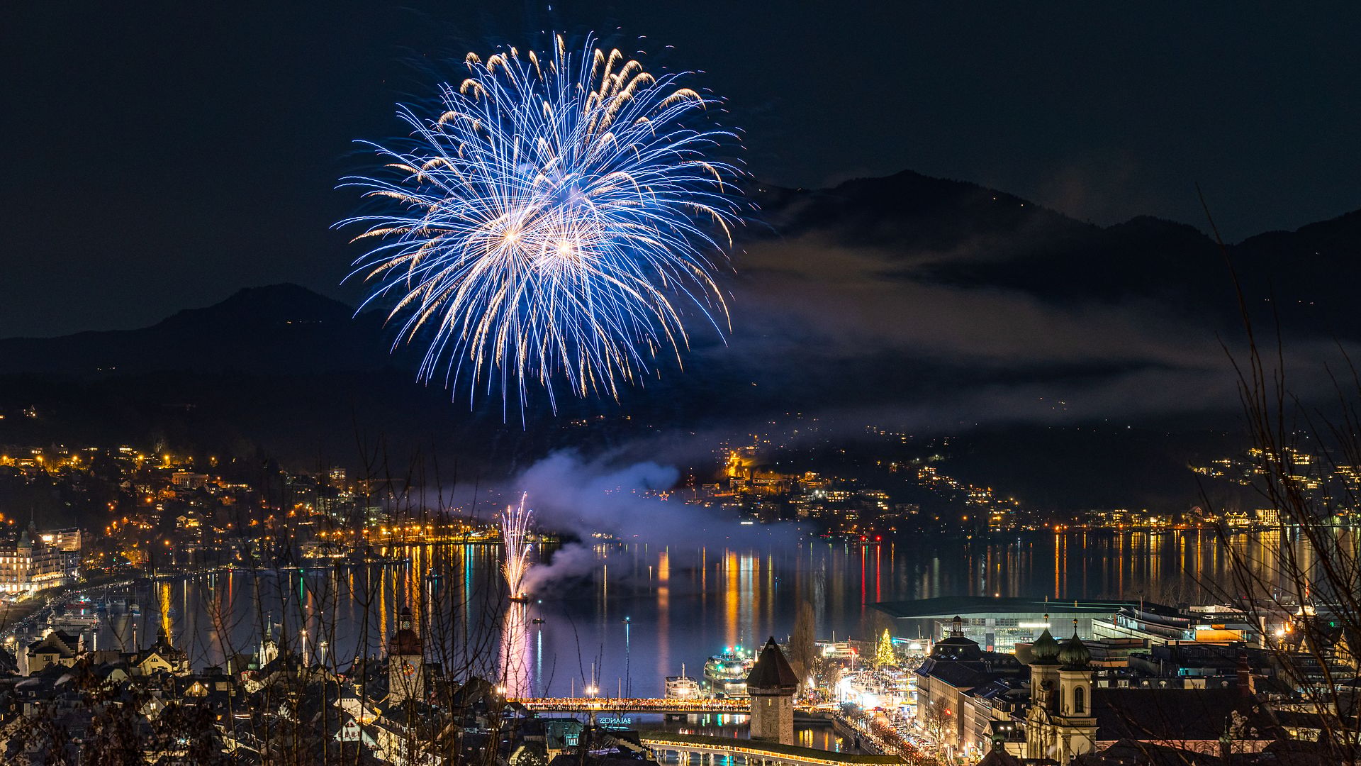 Feuerwerk über Luzern am Vierwaldstättersee bei Nacht mit beleuchteter Stadt und Bergkulisse.