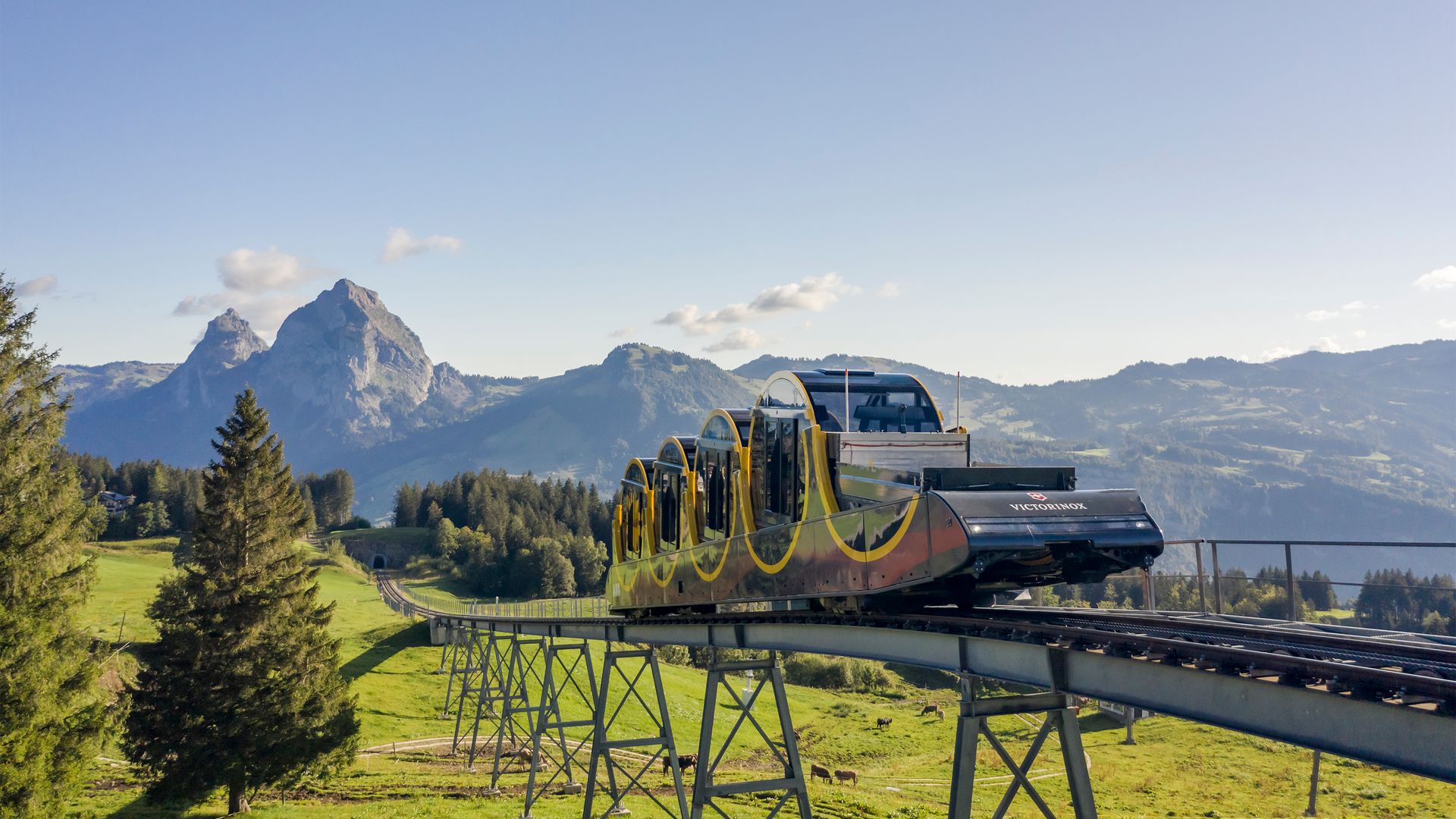 Stoos funicular travels on an elevated track through a sunny mountain landscape with views of distinctive peaks.