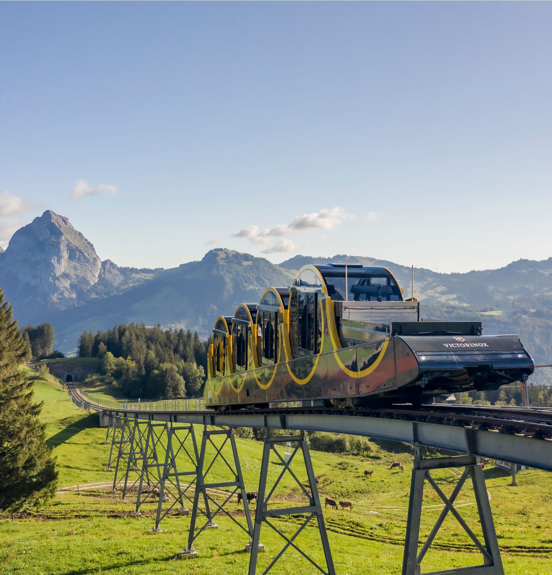 Stoos funicular travels on an elevated track through a sunny mountain landscape with views of distinctive peaks.
