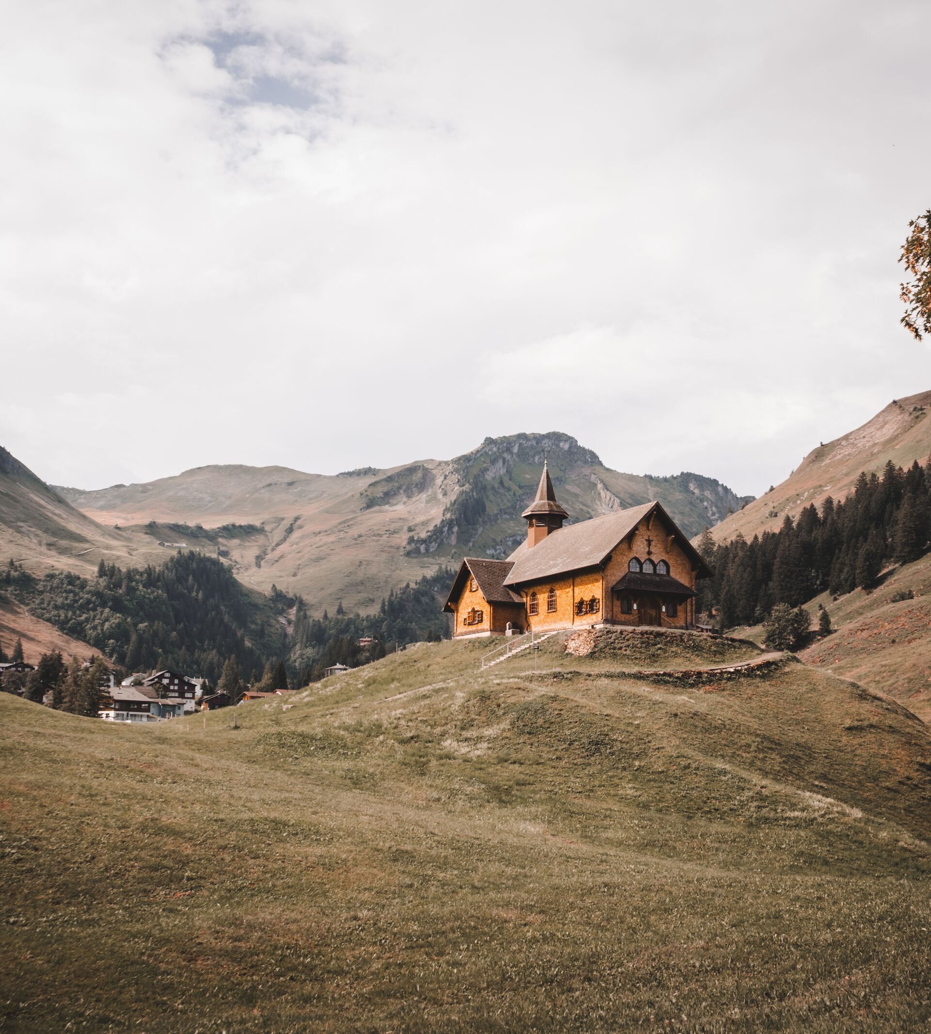 Kapelle auf einem Hügel in alpiner Landschaft, umgeben von Wiesen und Bergen.