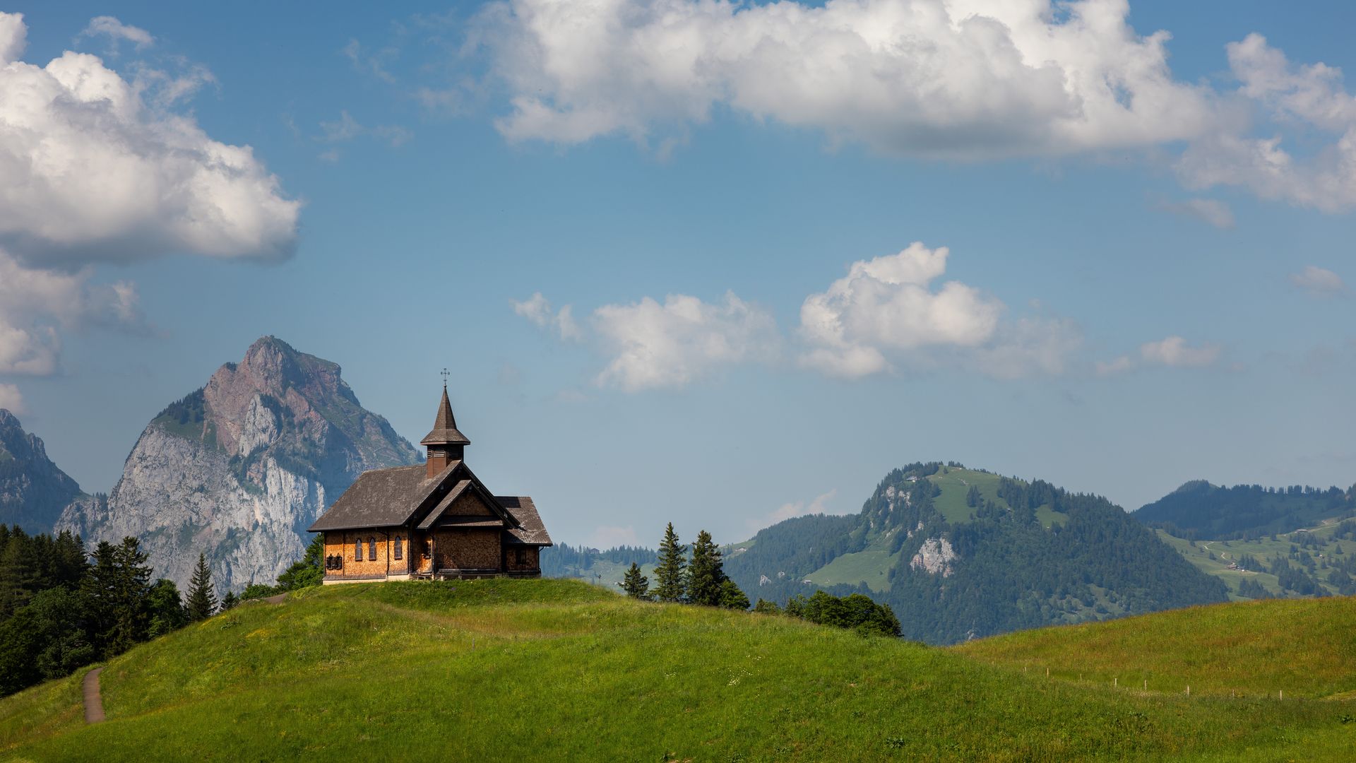 Kapelle auf einem grünen Hügel mit Blick auf markante Berggipfel und blauen Himmel.