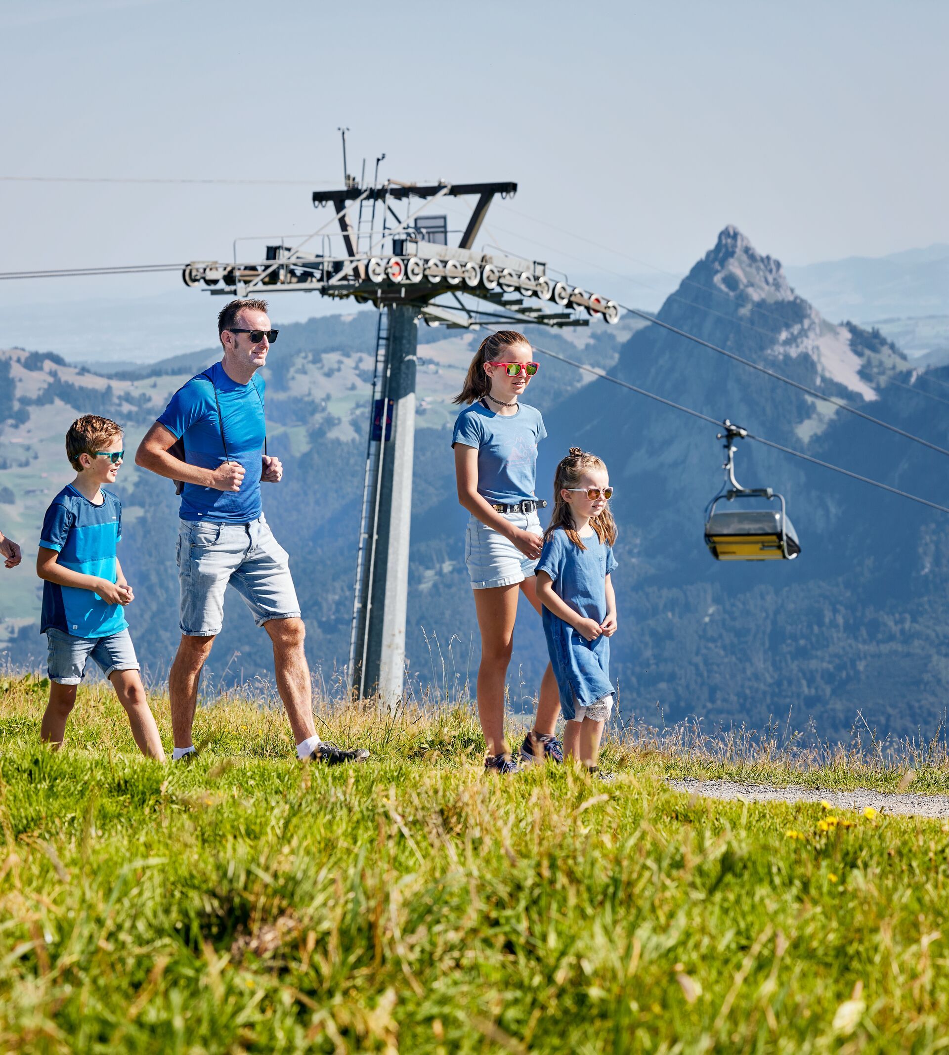Familie wandert über eine sonnige Bergwiese, im Hintergrund eine Sesselbahn und markante Gipfel.