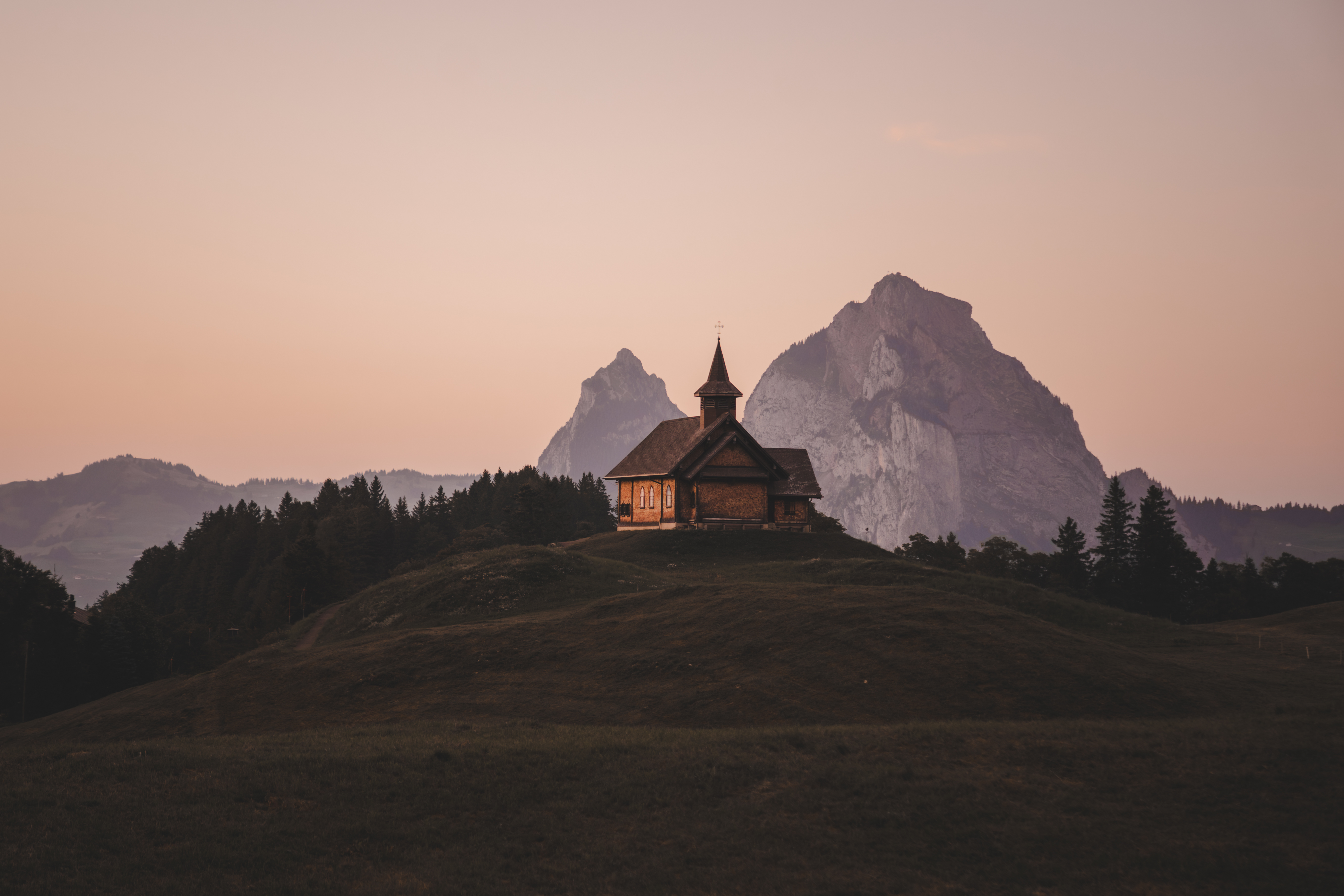 Kapelle auf einem Hügel im Abendlicht mit Berggipfeln im Hintergrund.