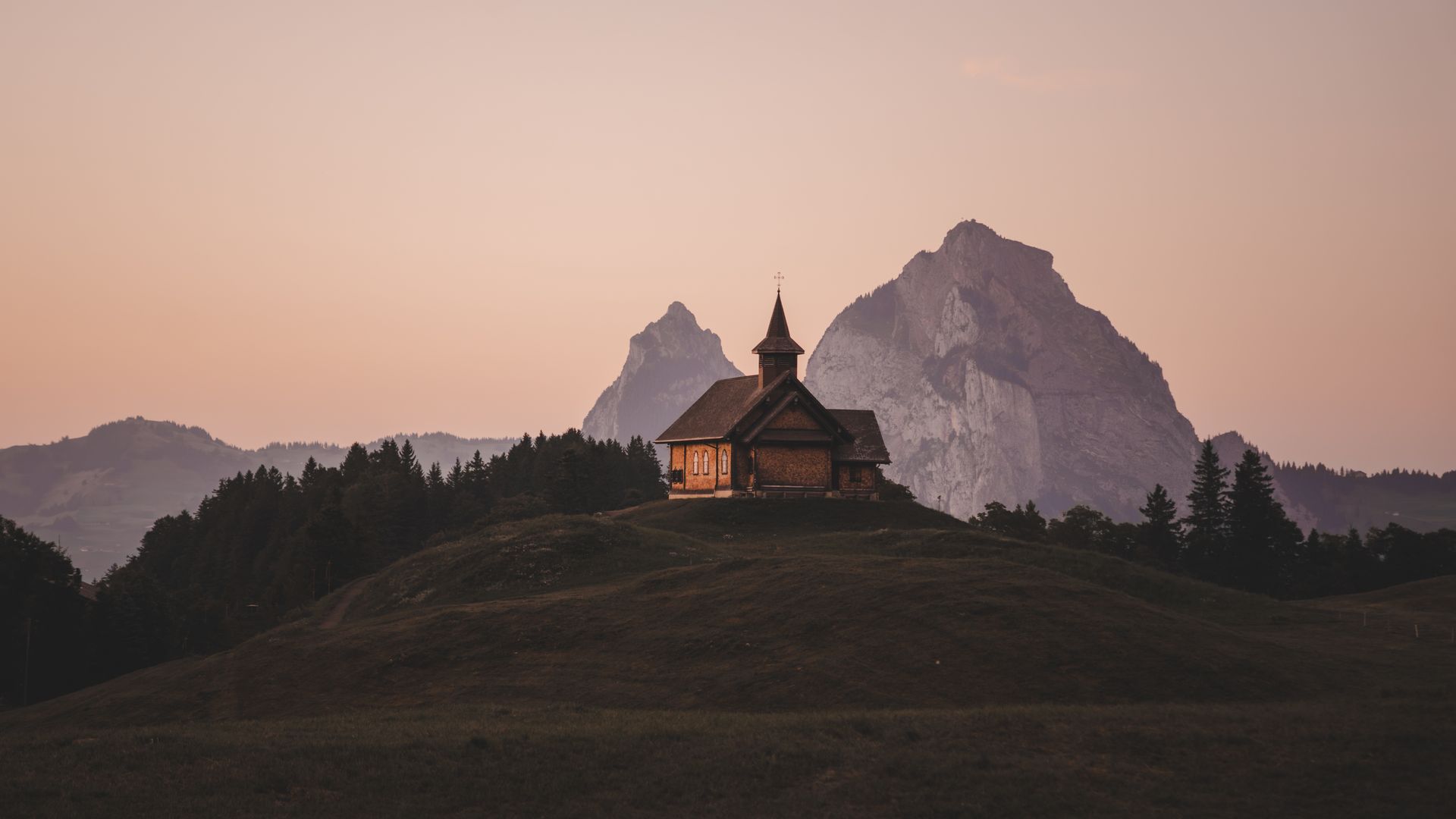 Kapelle auf einem Hügel im Abendlicht mit Berggipfeln im Hintergrund.