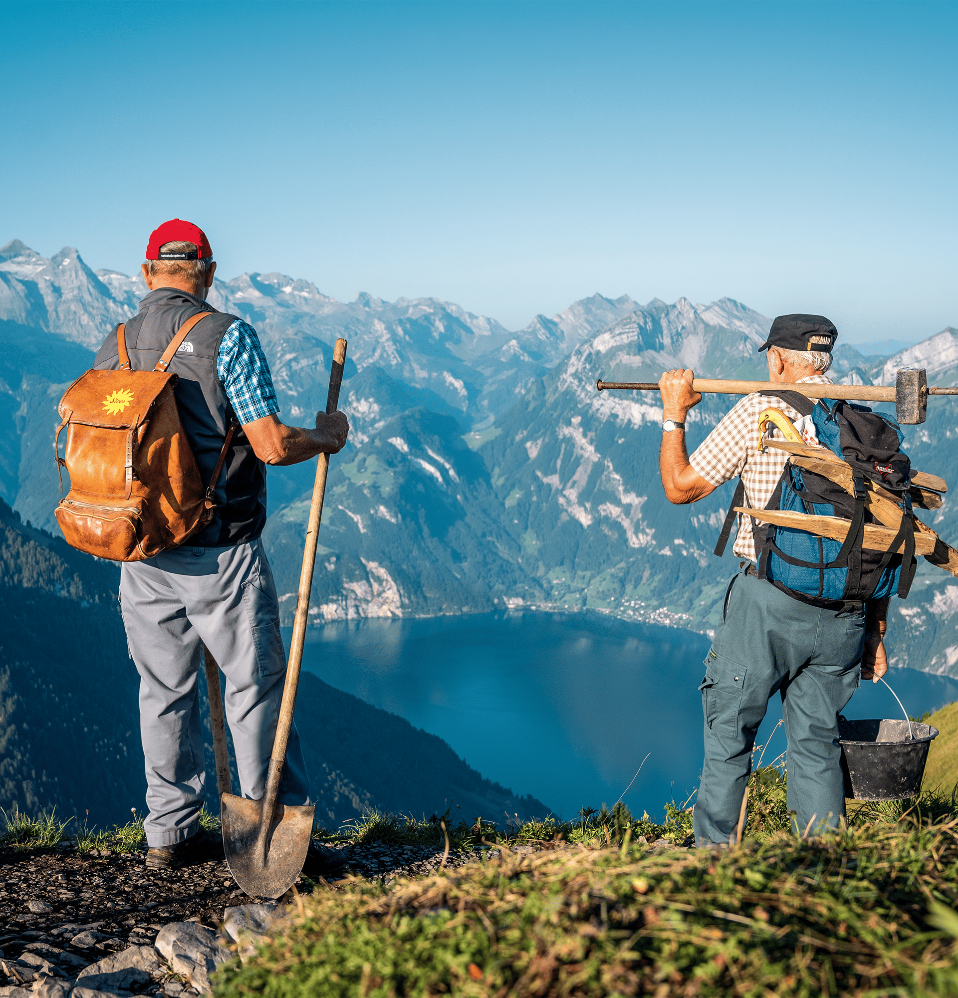 Two hikers standing on a mountain ridge with backpacks overlooking the surrounding Alps.