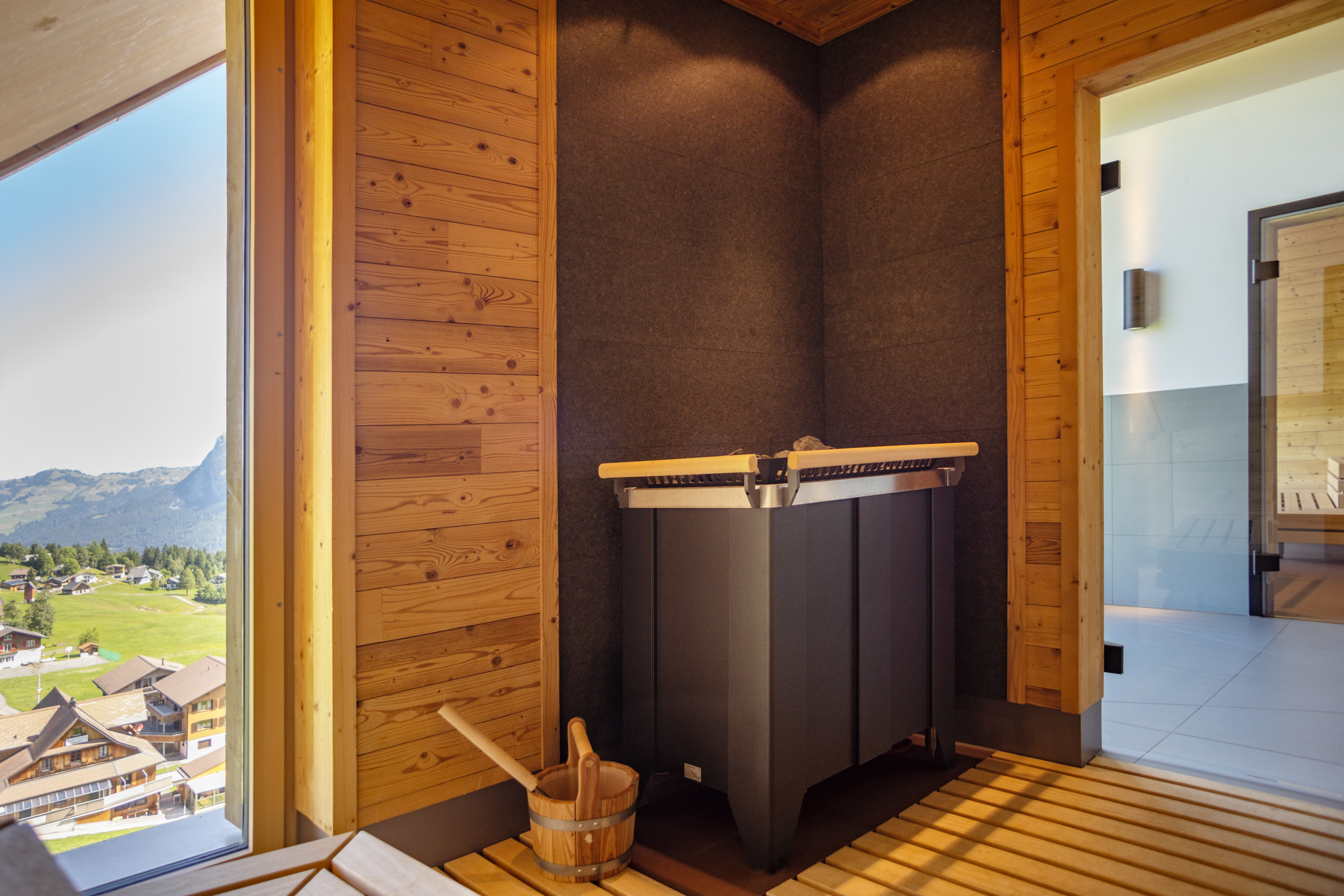 Moderner Saunabereich aus Holz mit Ofen und Blick durch grosse Fenster in die Berglandschaft.