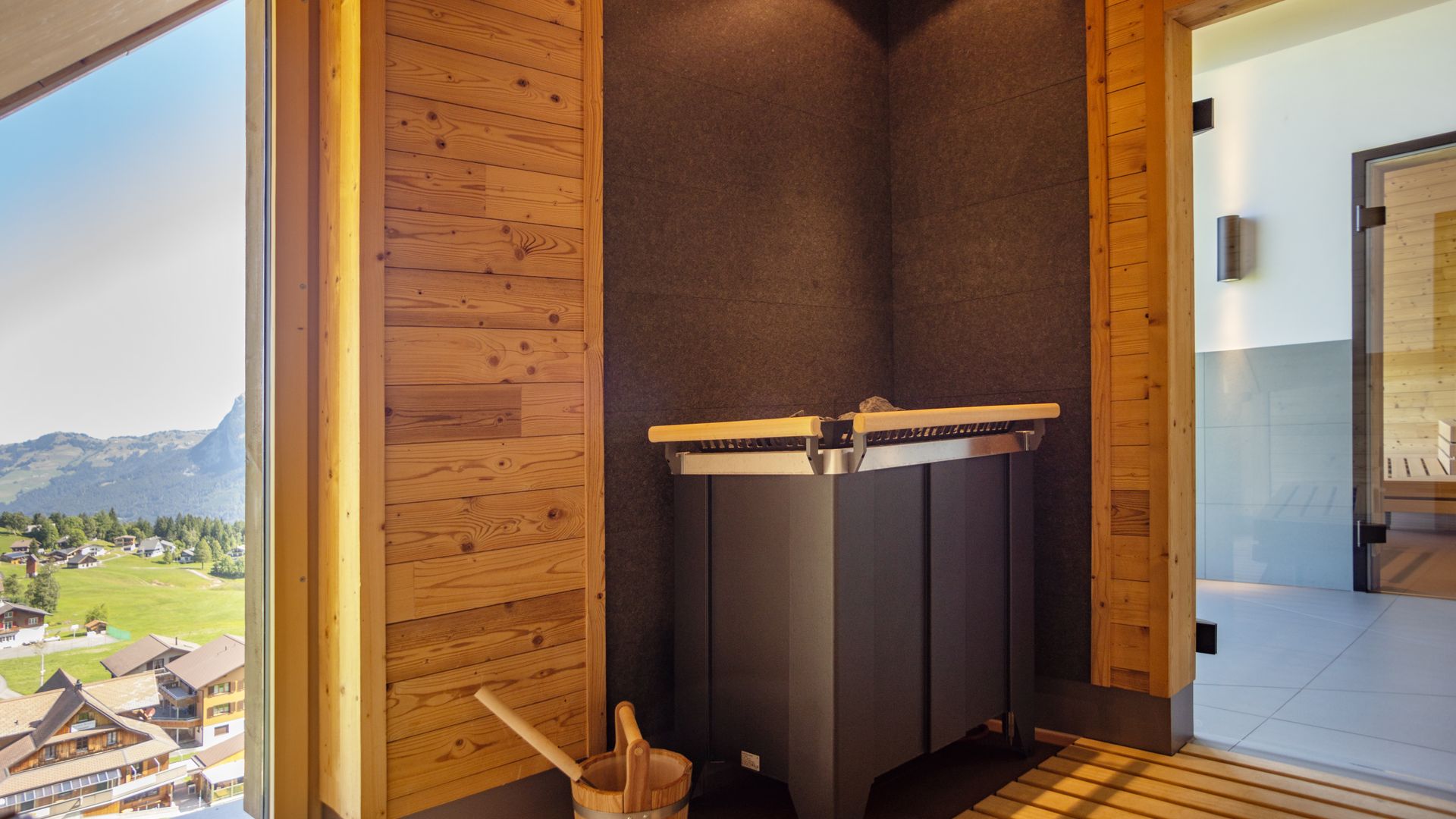 Moderner Saunabereich aus Holz mit Ofen und Blick durch grosse Fenster in die Berglandschaft.