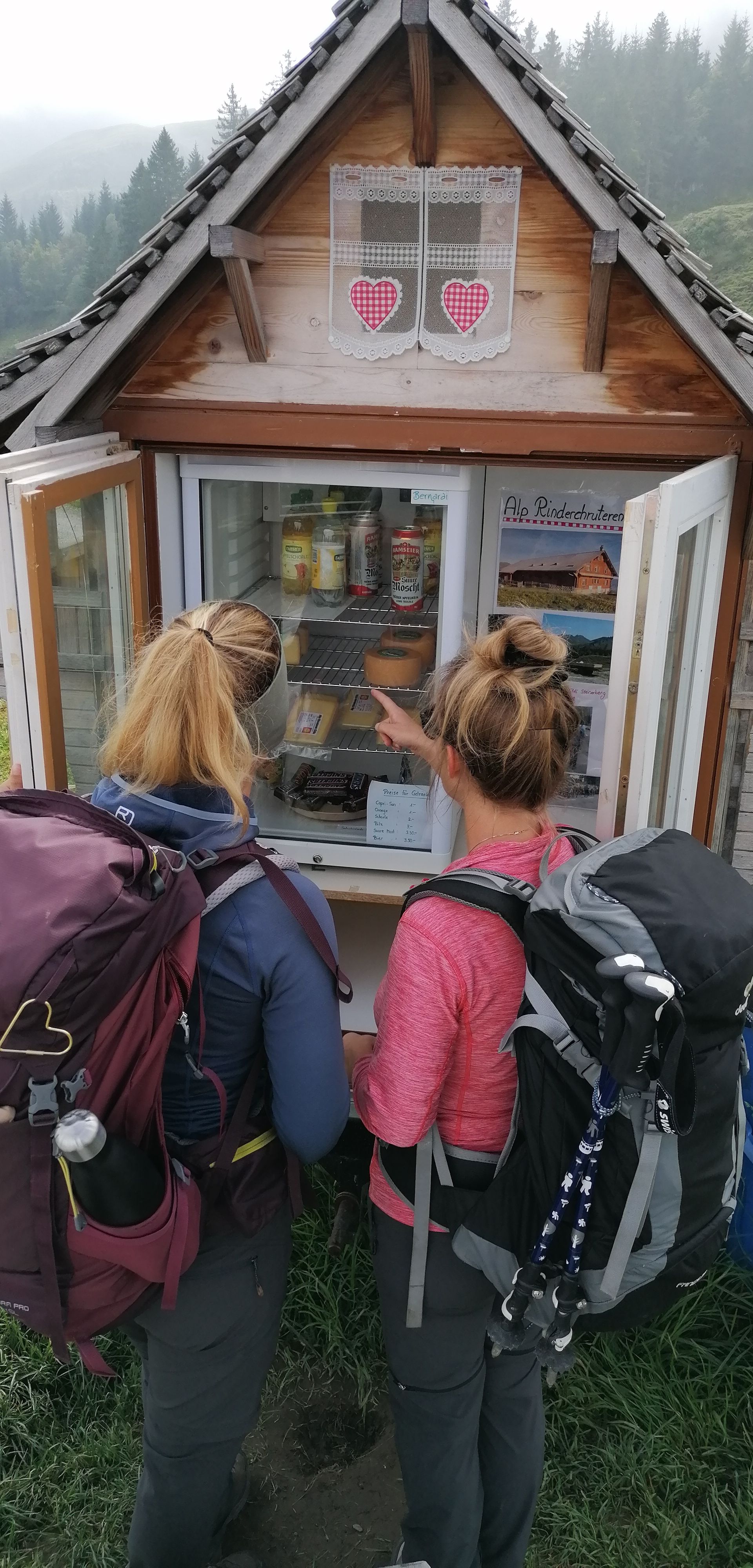 Two hikers stand in front of a small self-service hut with products in an alpine setting.