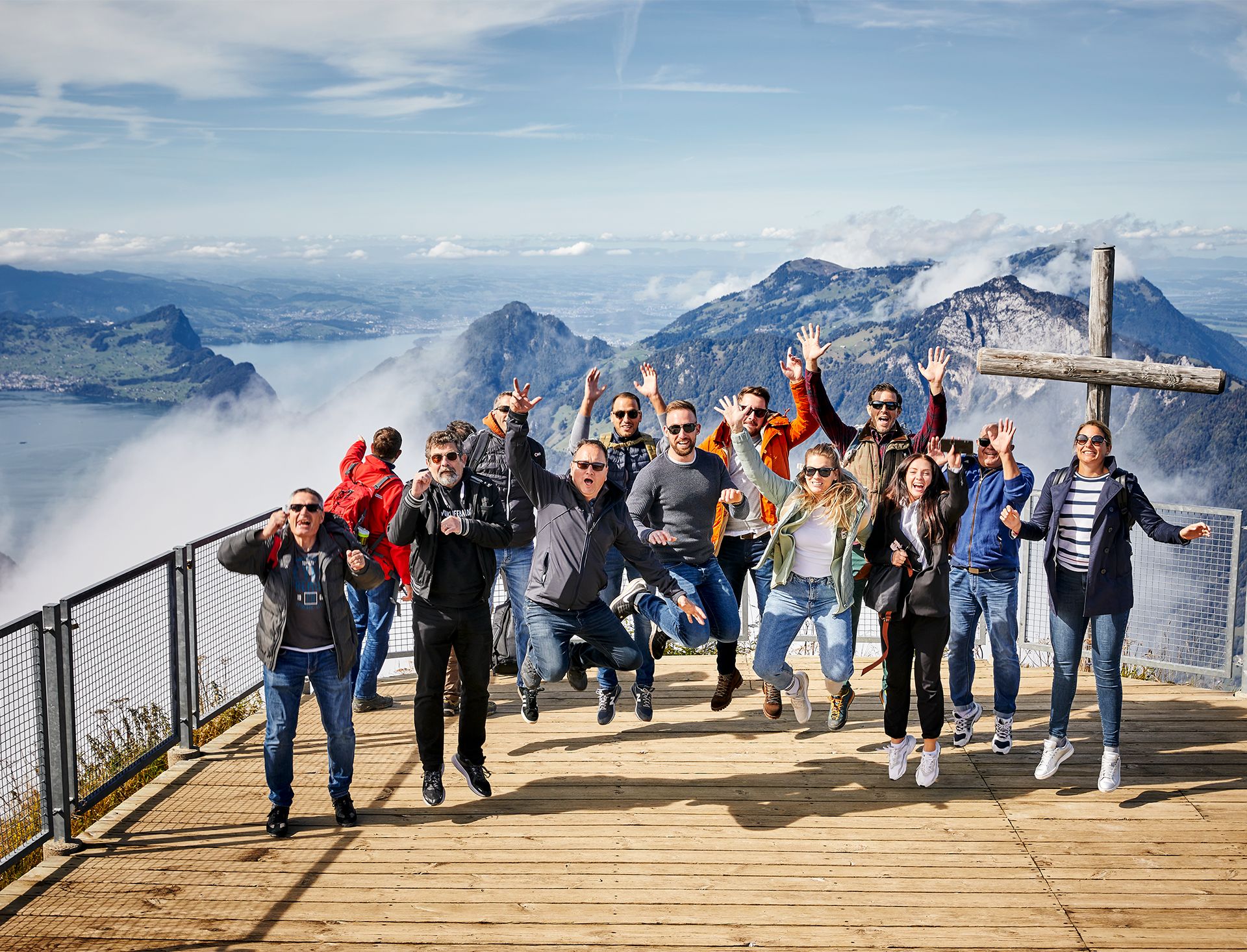 A group of people jumps and celebrates on a mountain viewing platform next to a large wooden cross, overlooking a lake and peaks.