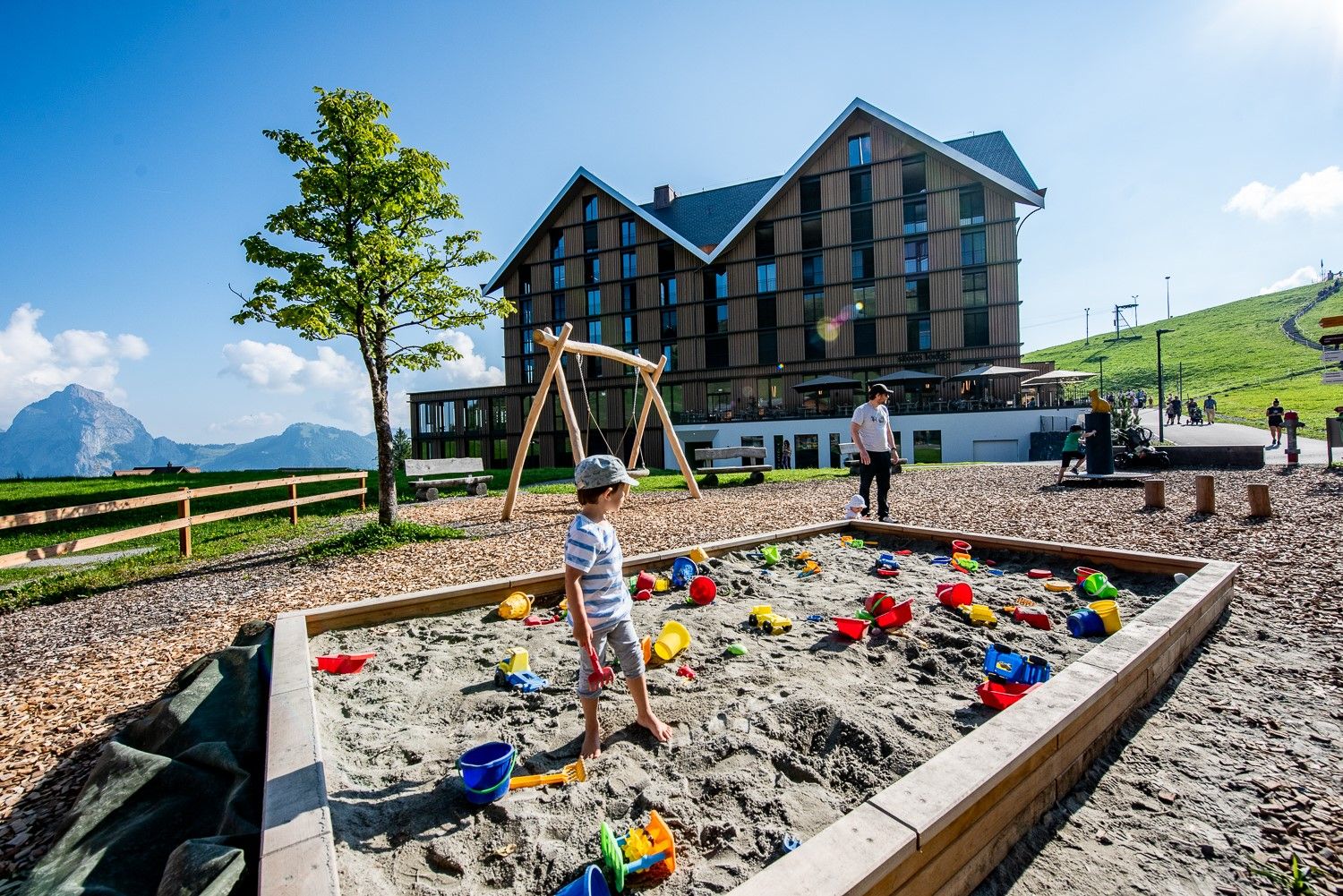 Sandspielplatz mit Spielzeug vor einem Gebäude auf einer Bergwiese mit Blick auf die Alpen.