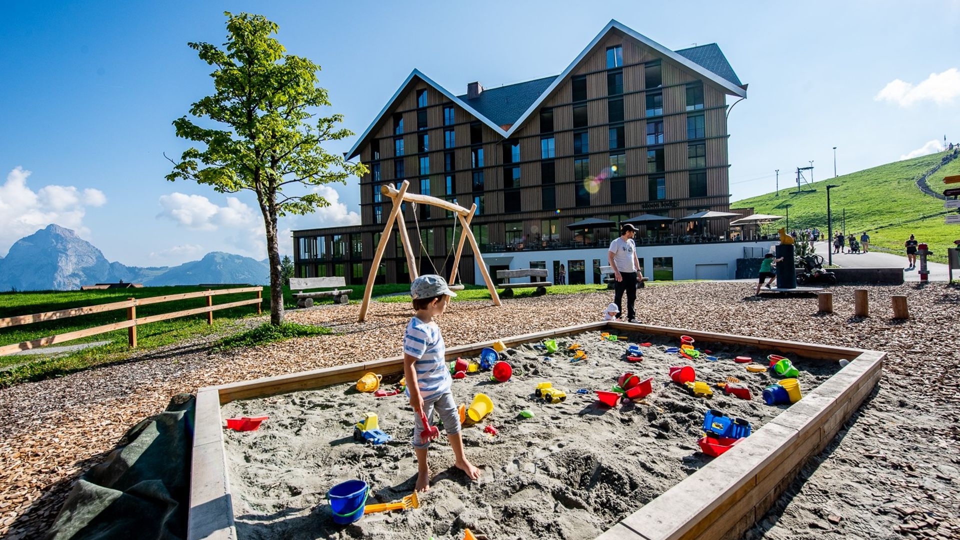 Sand playground with toys in front of a building on a mountain meadow with views of the Alps.