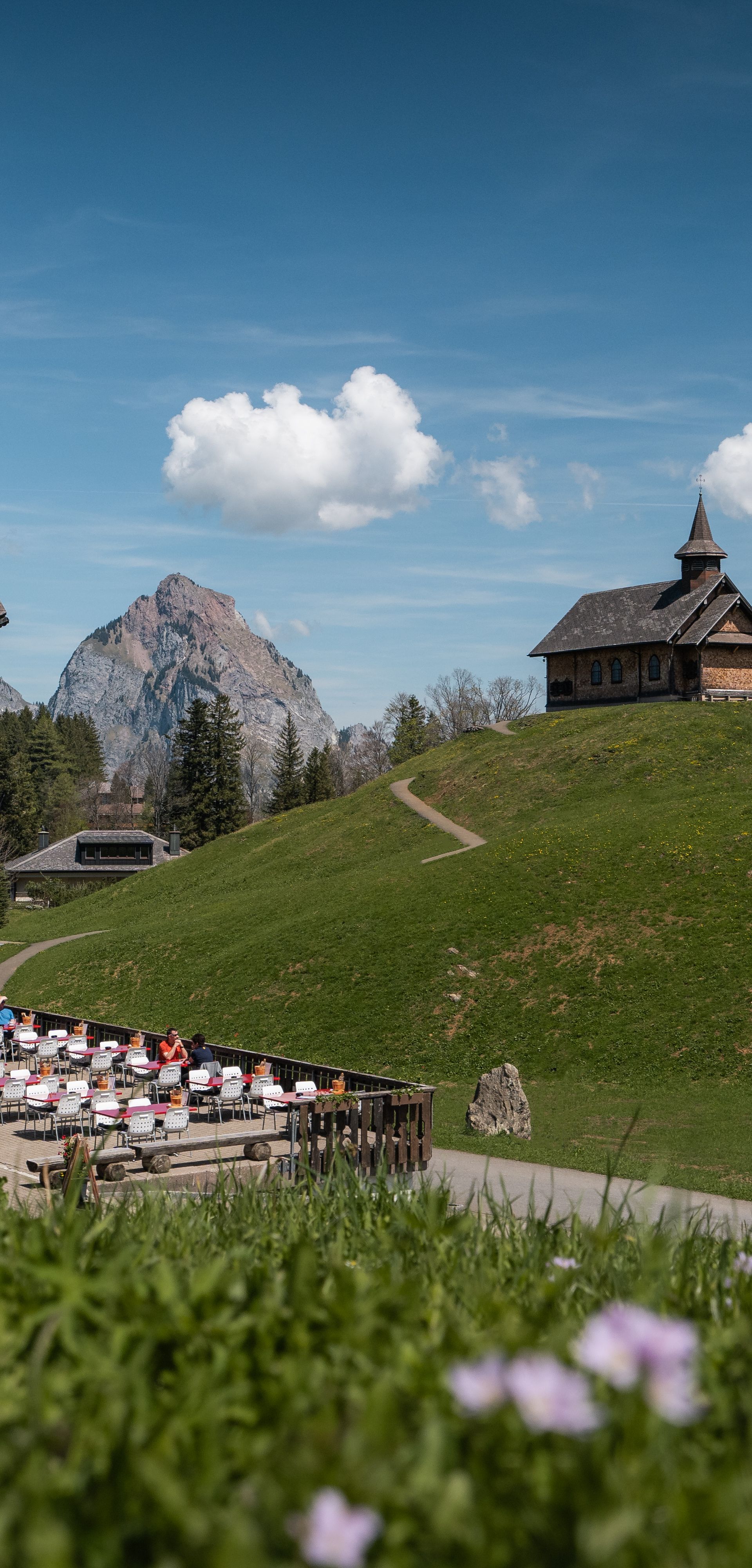 Bergrestaurant auf einer Wiese mit Terrasse, im Hintergrund eine Kapelle auf einem Hügel.