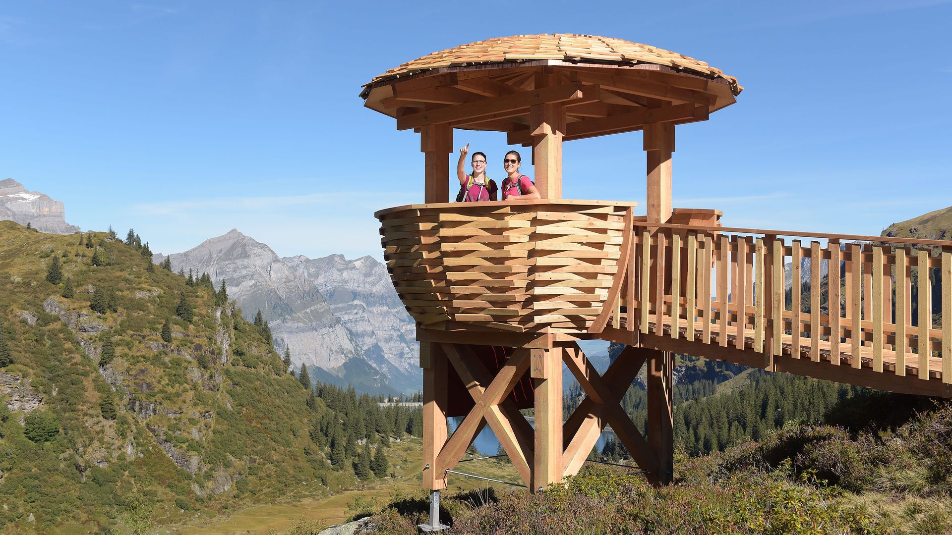 Two women stand on a wooden viewing platform on Mettmenalp and discuss their hiking route.