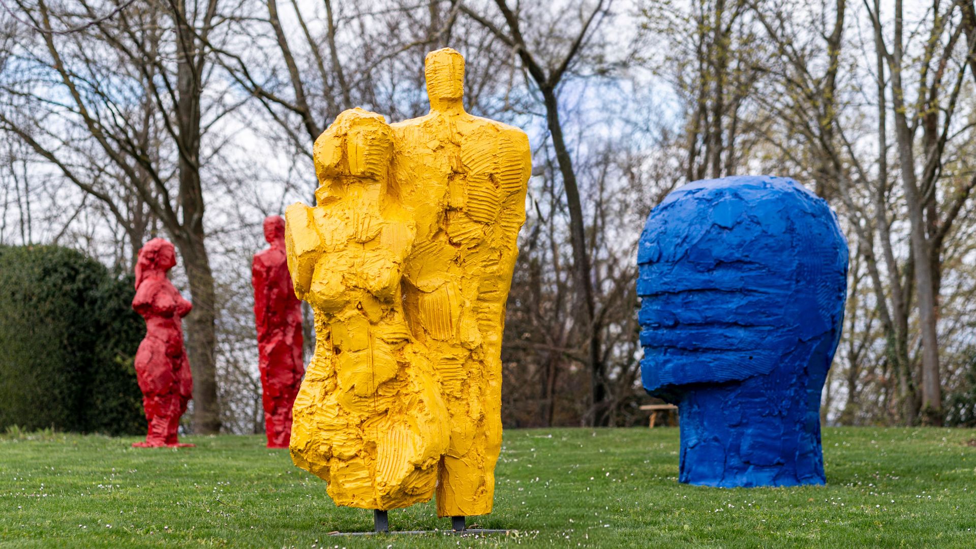 Mehrere grob modellierte, farbige Skulpturen in menschlicher Form auf einer Wiese vor kahlen Bäumen.