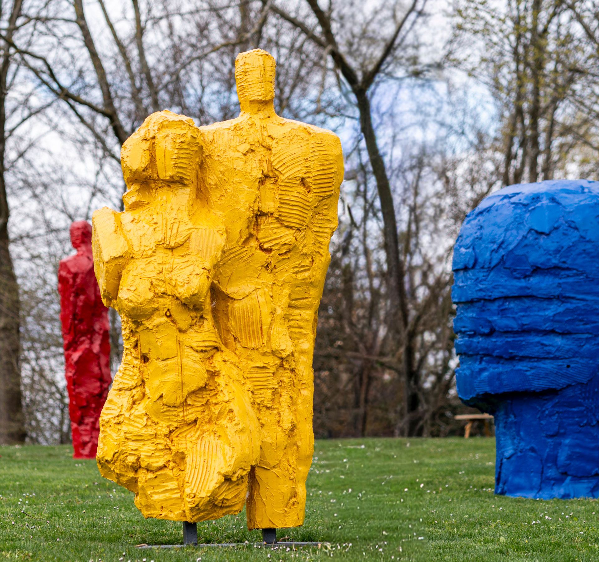 Diverse sculture colorate a forma umana grossolanamente modellate su un prato davanti ad alberi spogli.