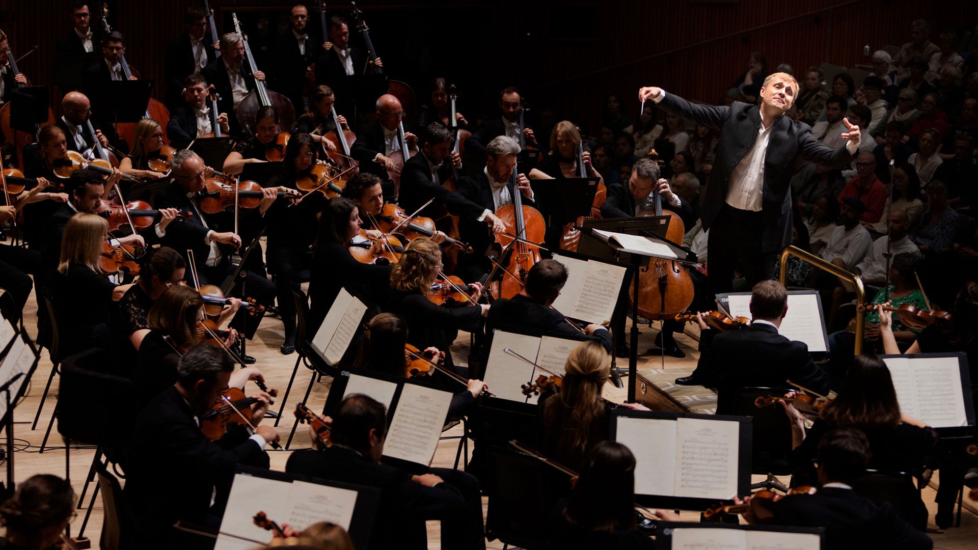 Dirigent vor einem Orchester mit Streichern und anderen Musikern in einem Konzertsaal.