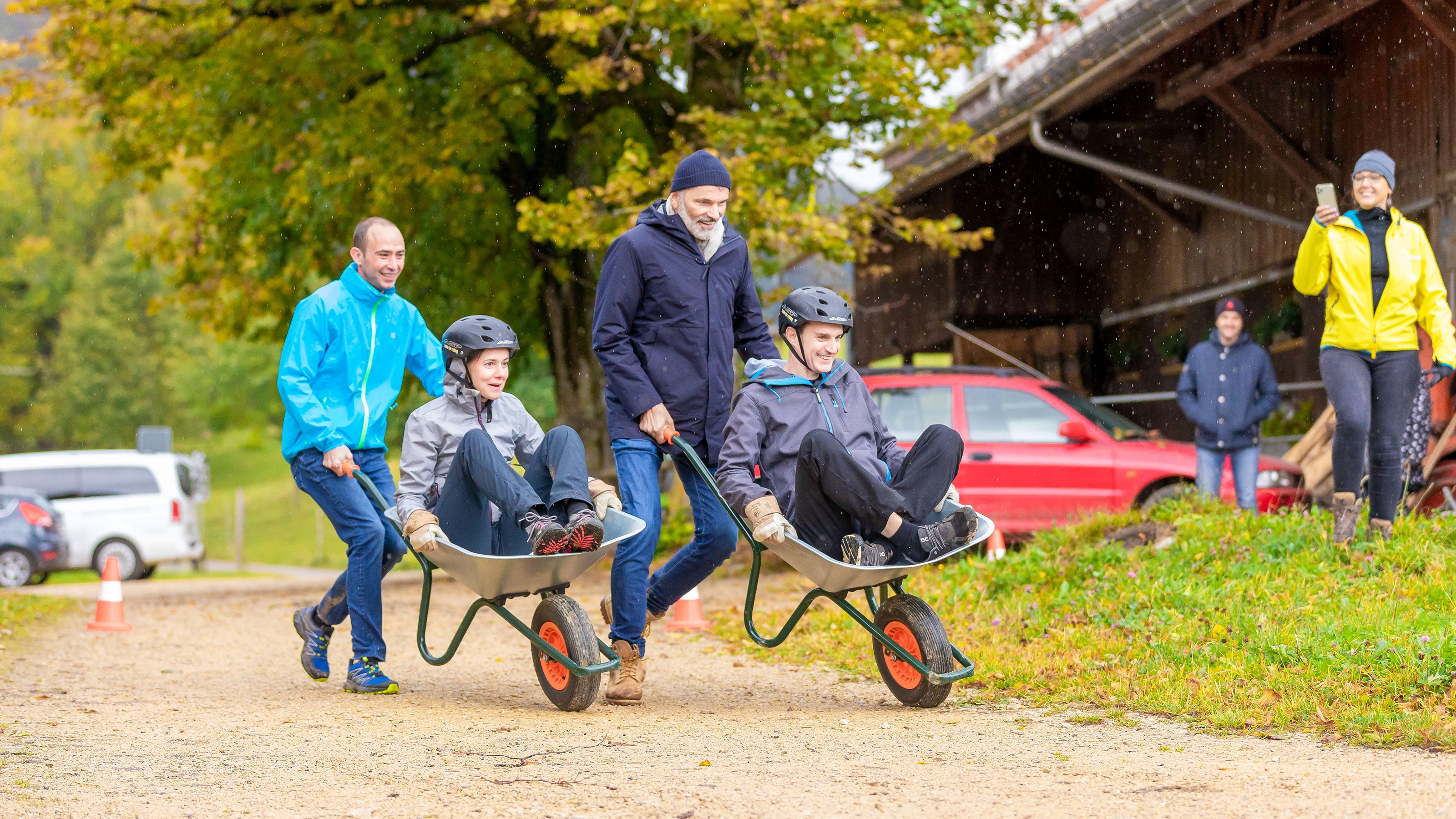 Two men are pushing a woman and a man in a wheelbarrow along a road that belongs to a farm. It's about which team is faster.