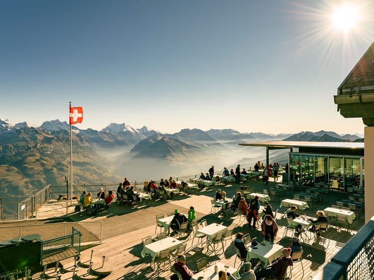 Eine Gruppe von Menschen sitzt an Tischen und Stühlen auf einem Dach mit Blick auf die Berge.