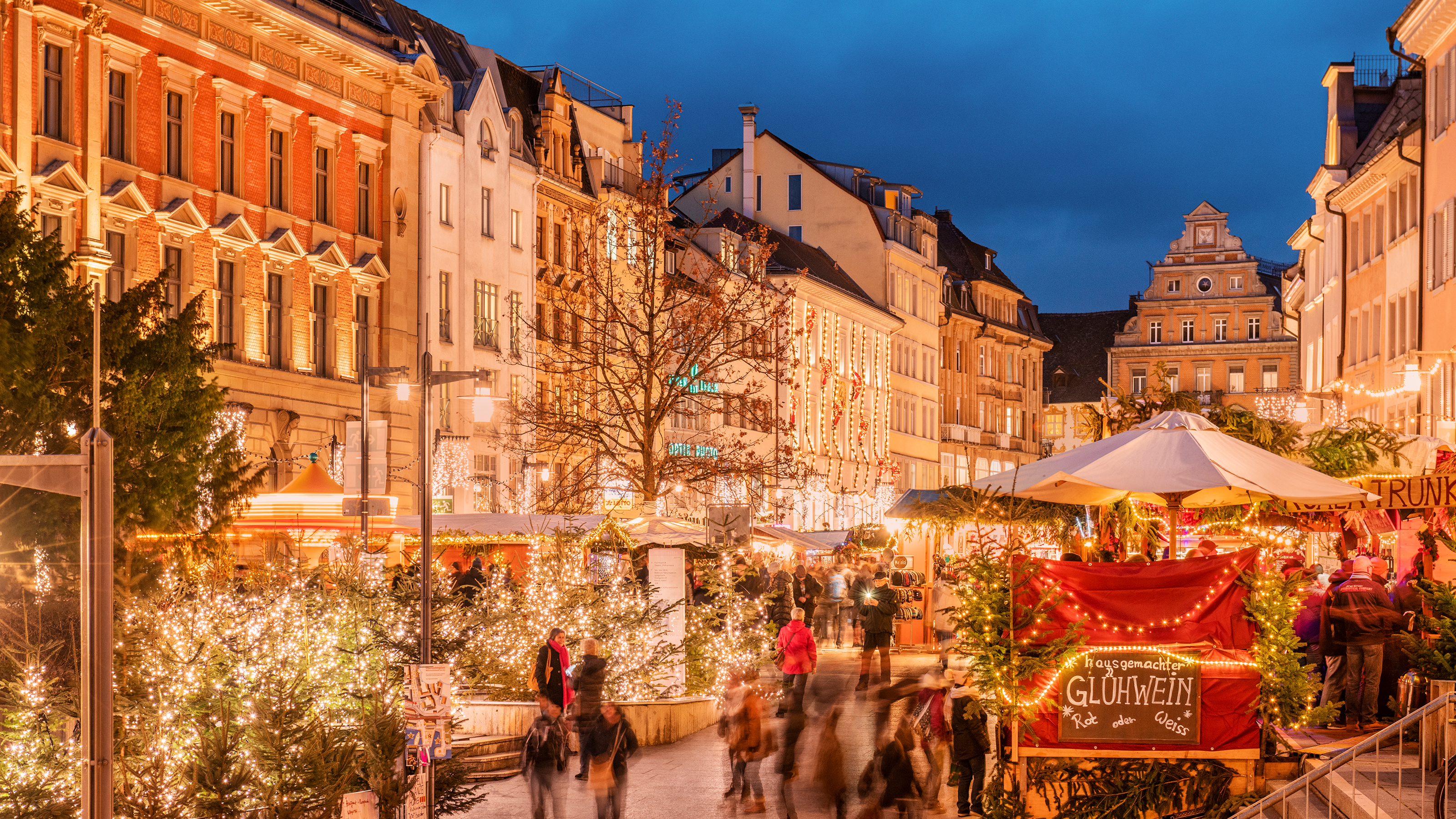 A busy outdoor Christmas market at dusk is decorated with twinkling lights.