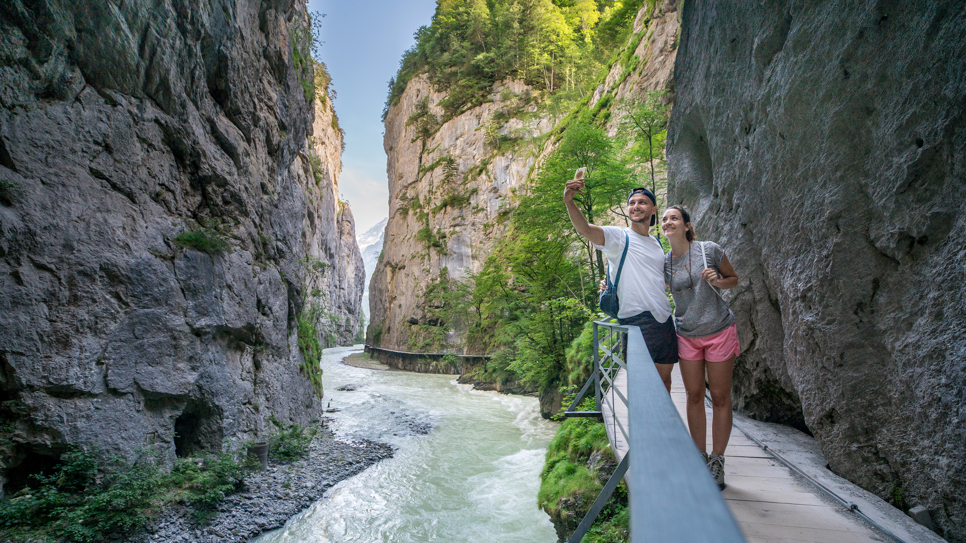 Two people take a selfie on a narrow walkway at the Aareschlucht with steep rock walls and a river.