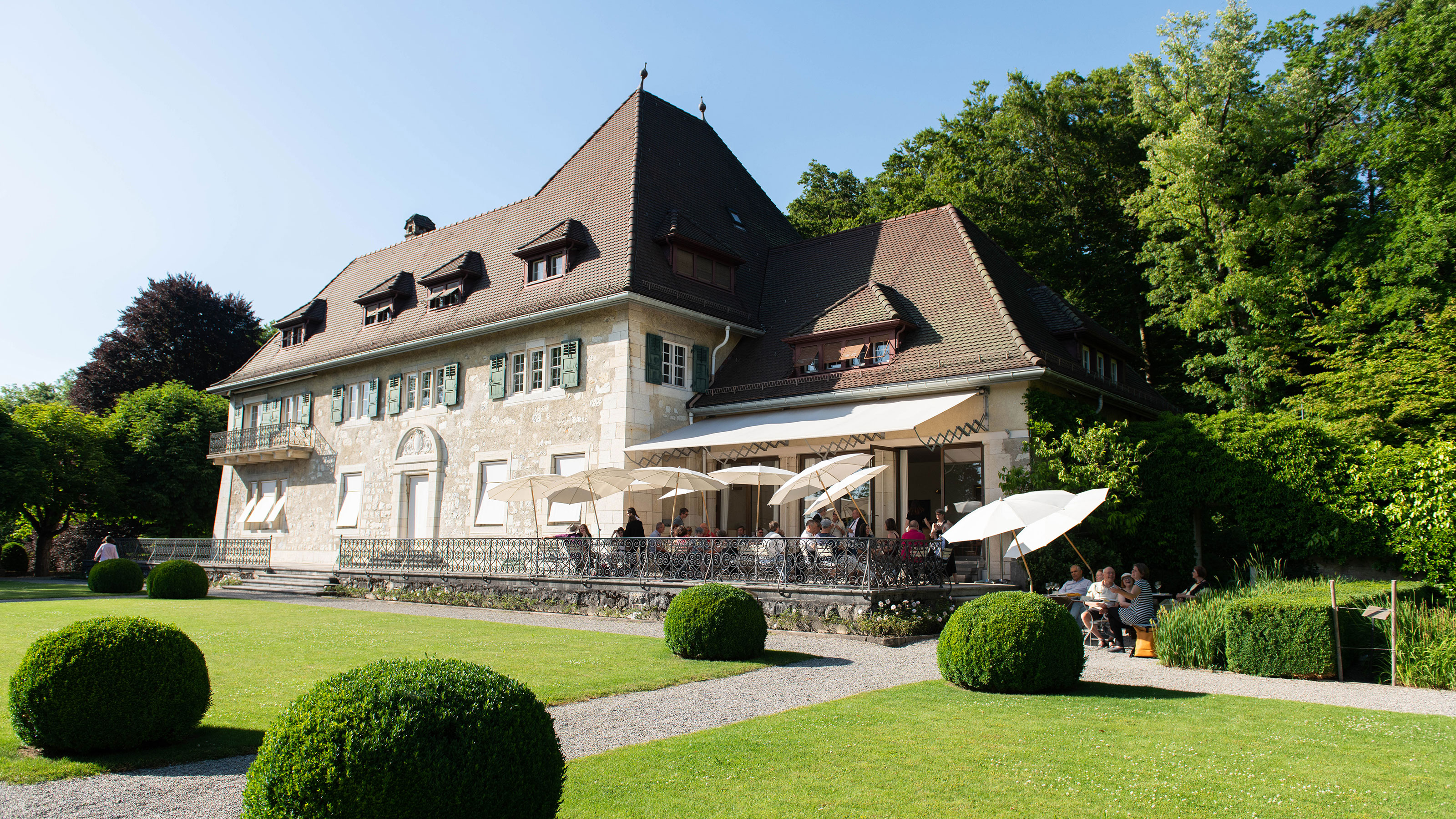 Large historic building with steep roof and terrace with umbrellas in front of manicured lawn and spherical bushes.